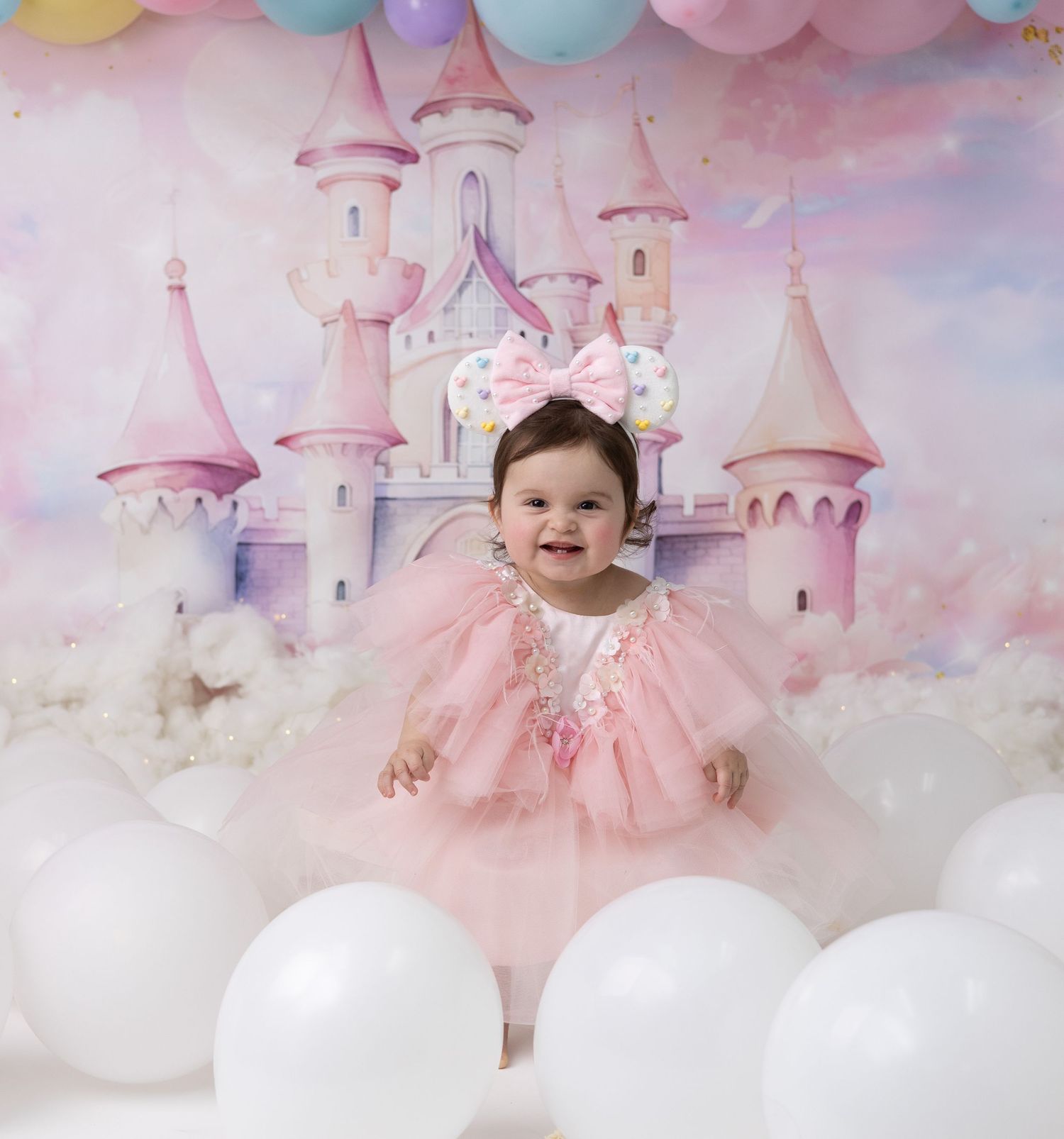 A one-year-old birthday girl in a Disney-themed outfit smiles surrounded by colorful decorations celebrating her magical first birthday