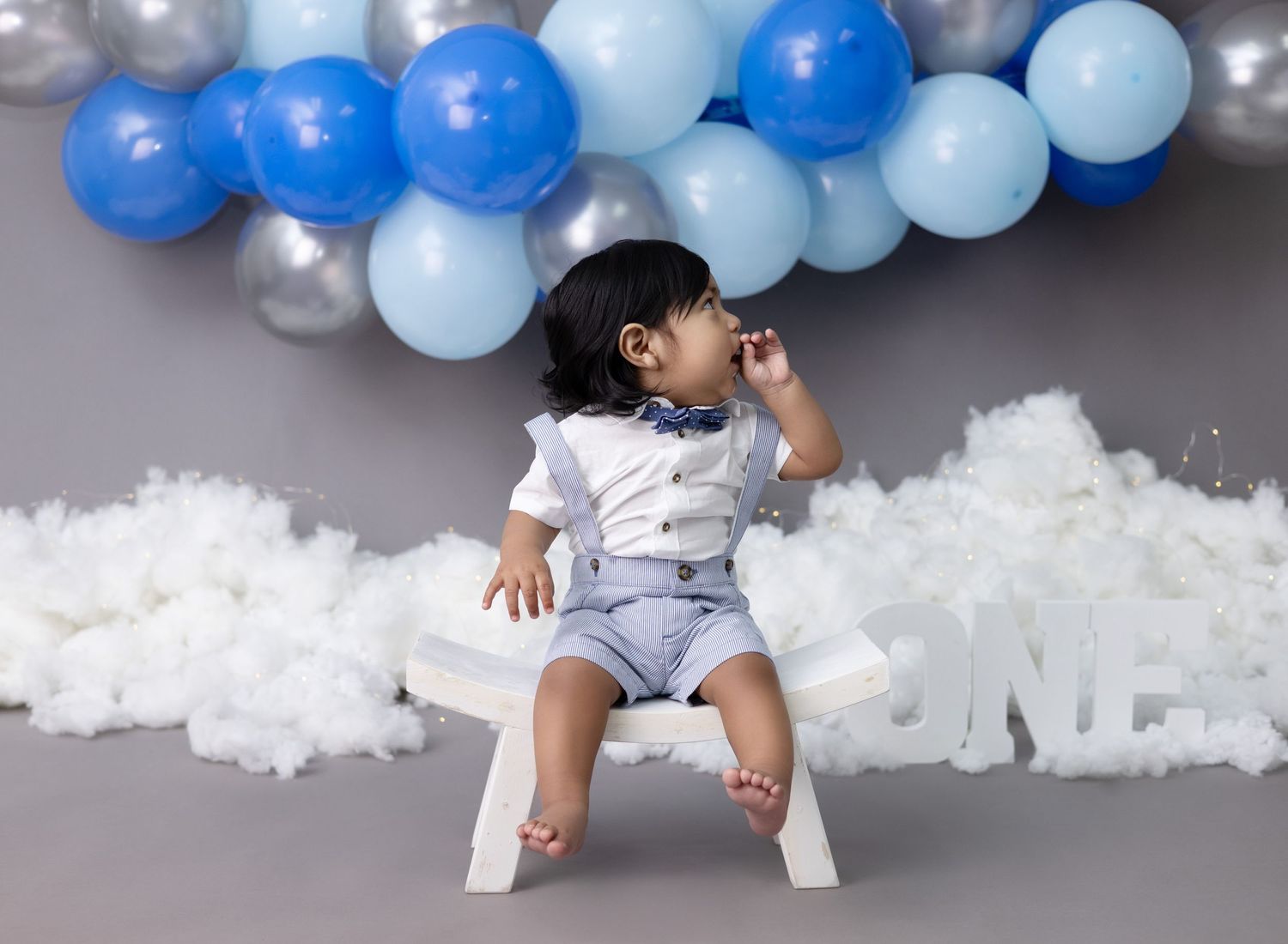 A one-year-old birthday boy sits on a chair, smiling happily during his first birthday photo session.