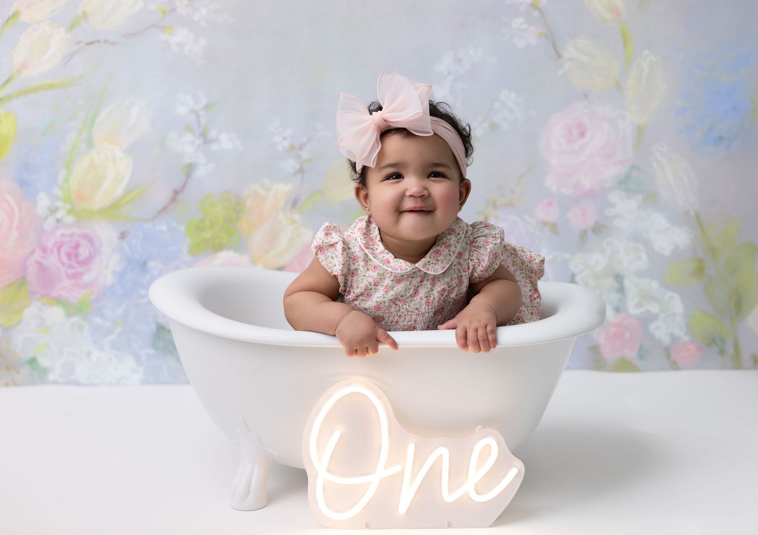 A birthday girl sits in a clawfoot bathtub, surrounded by a beautiful floral backdrop, smiling during her first birthday photoshoot.