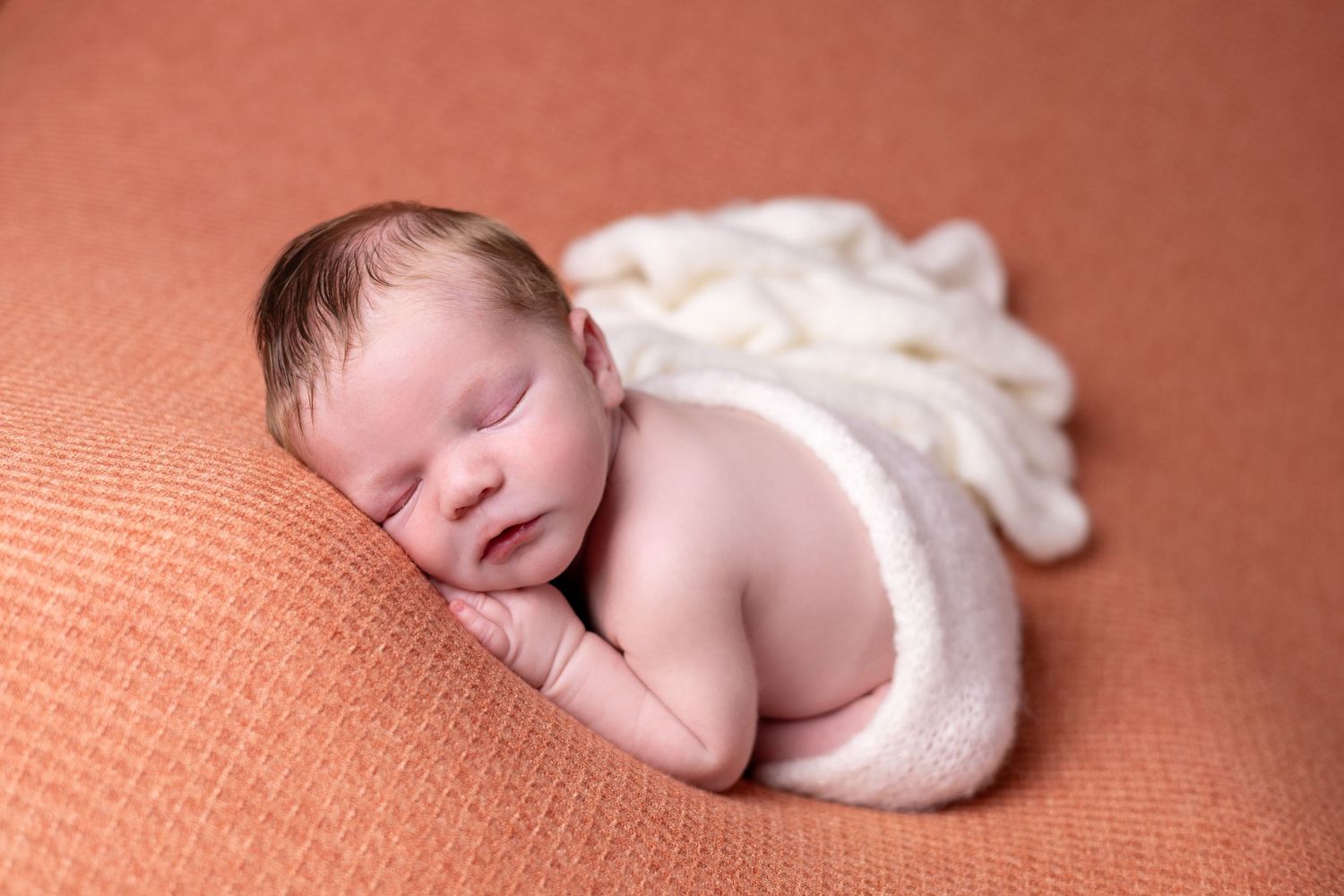 A newborn baby boy sleeps peacefully in the womb pose, swaddled and curled up.