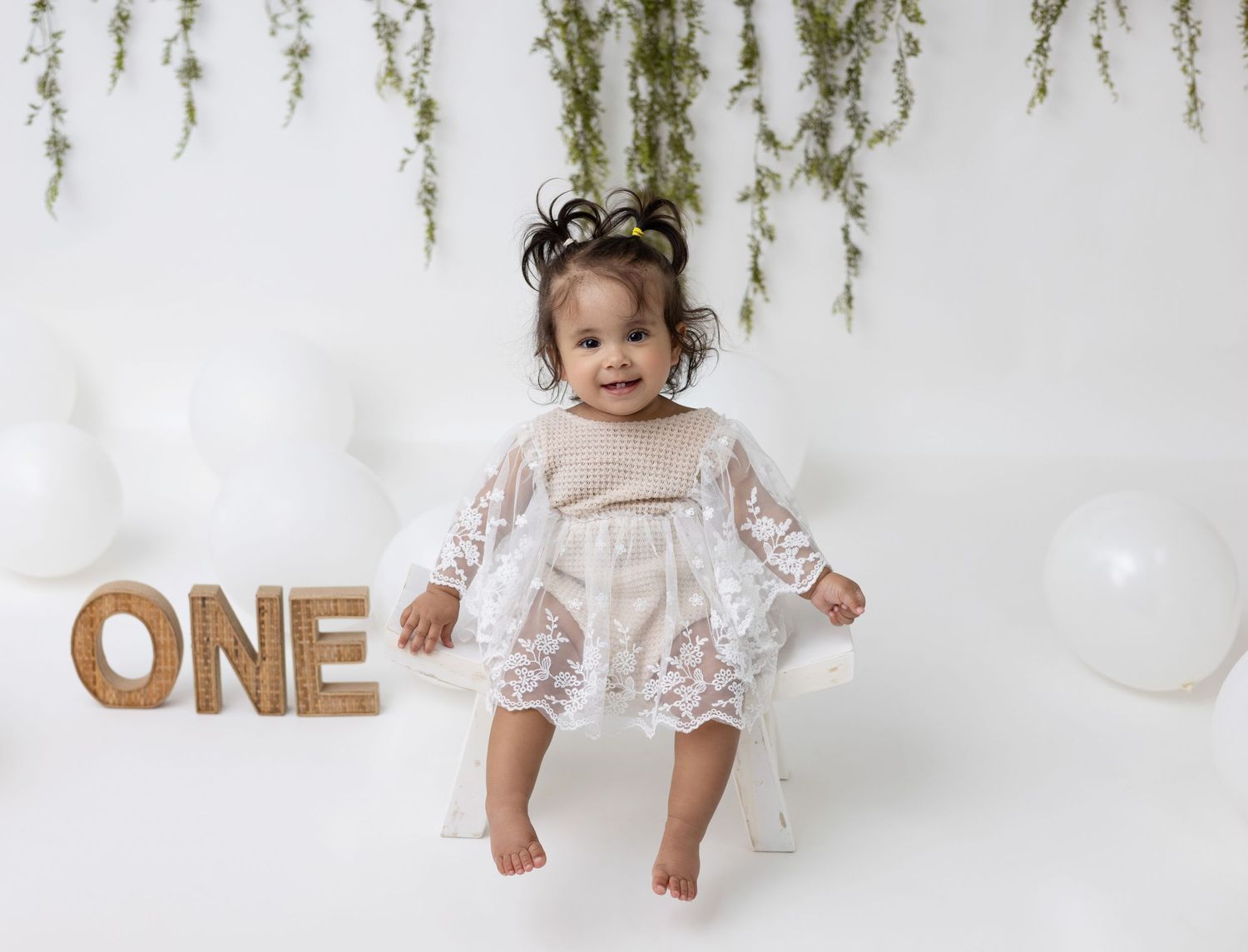 A one-year-old baby girl sits on a decorated set, wearing a cute birthday outfit and smiling joyfully.
