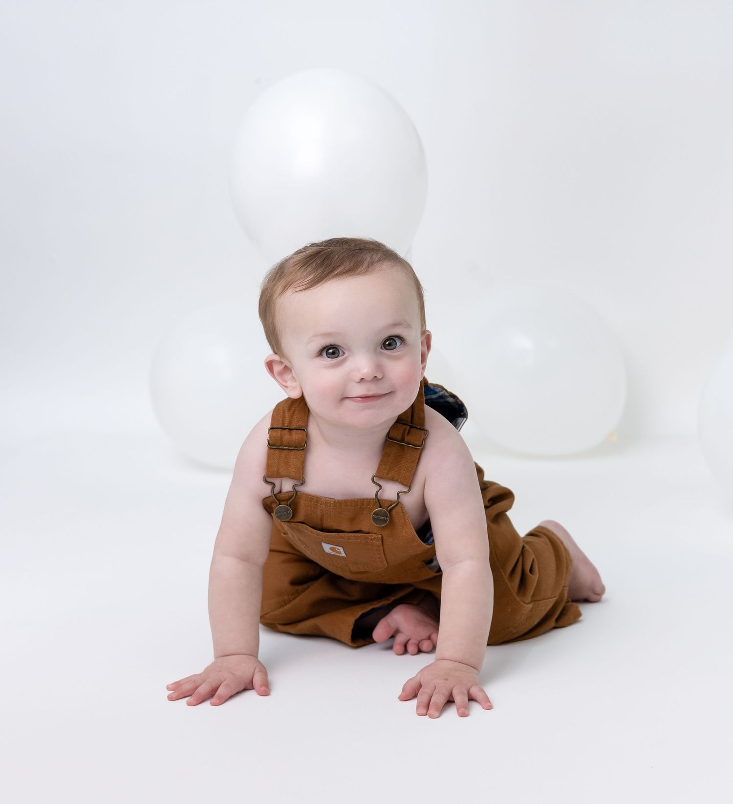 A crawling baby boy sits and smiles during his milestone session, surrounded by soft props and a cozy setup.