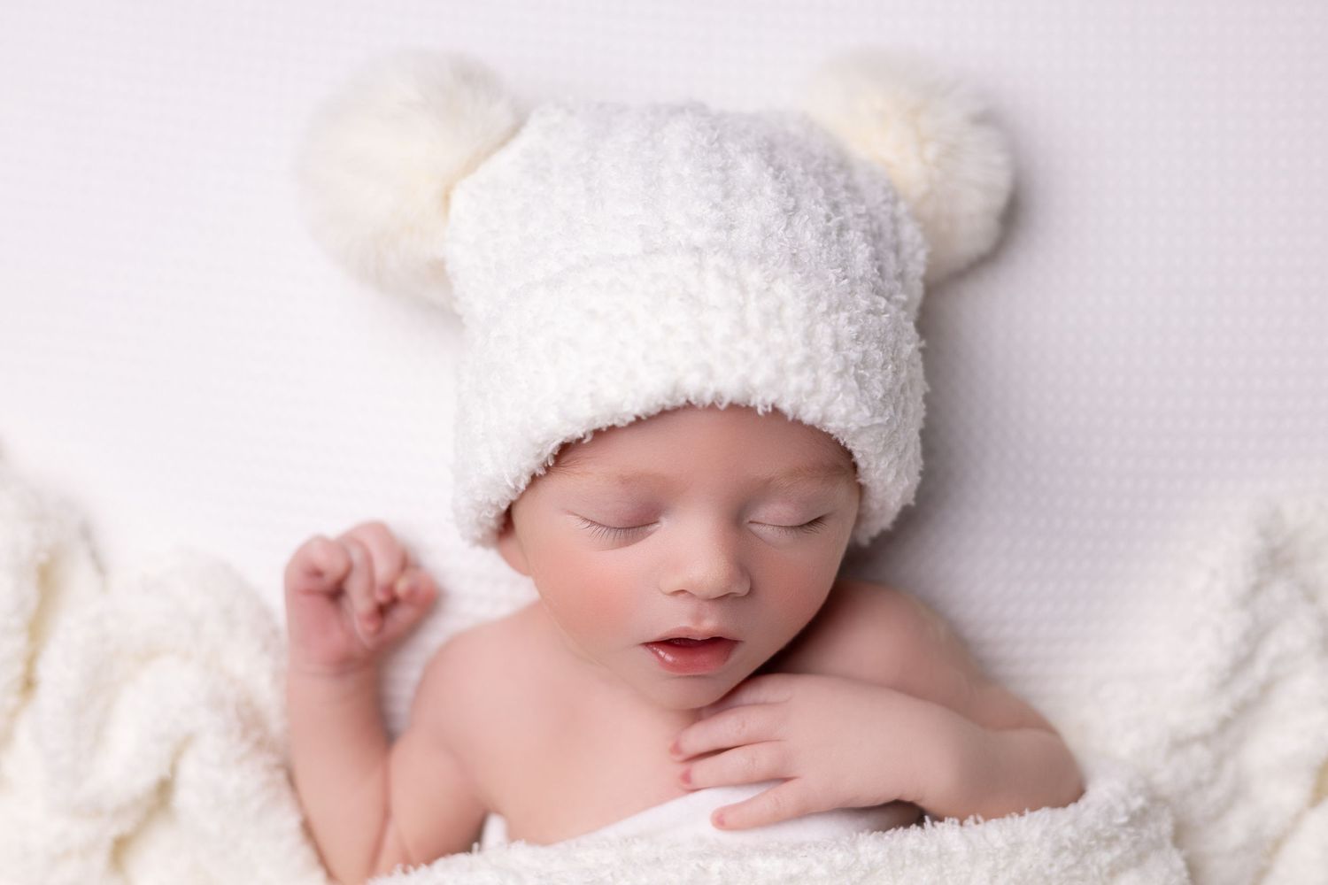 A newborn baby boy sleeps peacefully, swaddled in a soft white blanket, surrounded by an all-white backdrop.