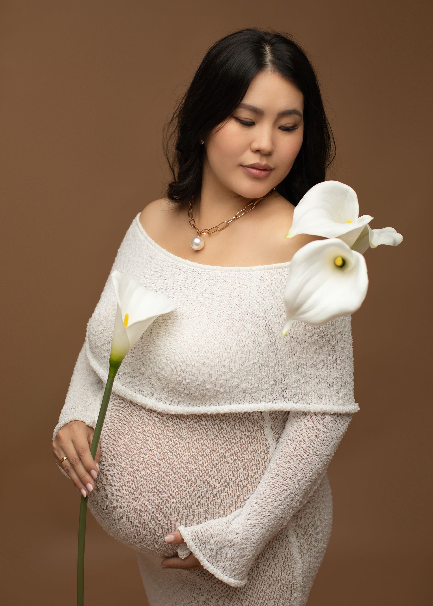 A pregnant woman in a white outfit poses in the studio, surrounded by soft lilies, creating an elegant and serene maternity session.