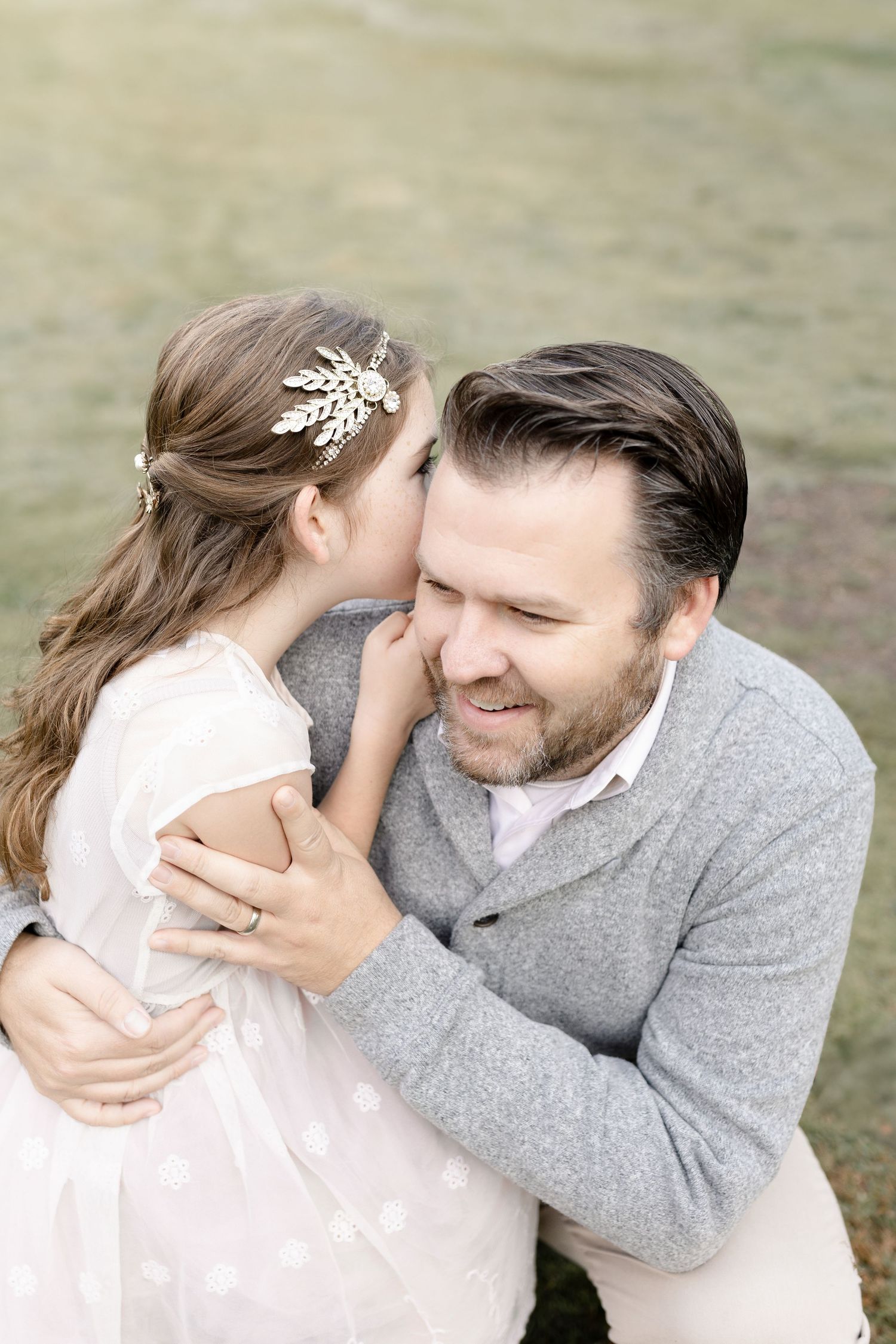 A little girl whispers in her dad's ear, sharing a sweet moment during an outdoor family photo session.