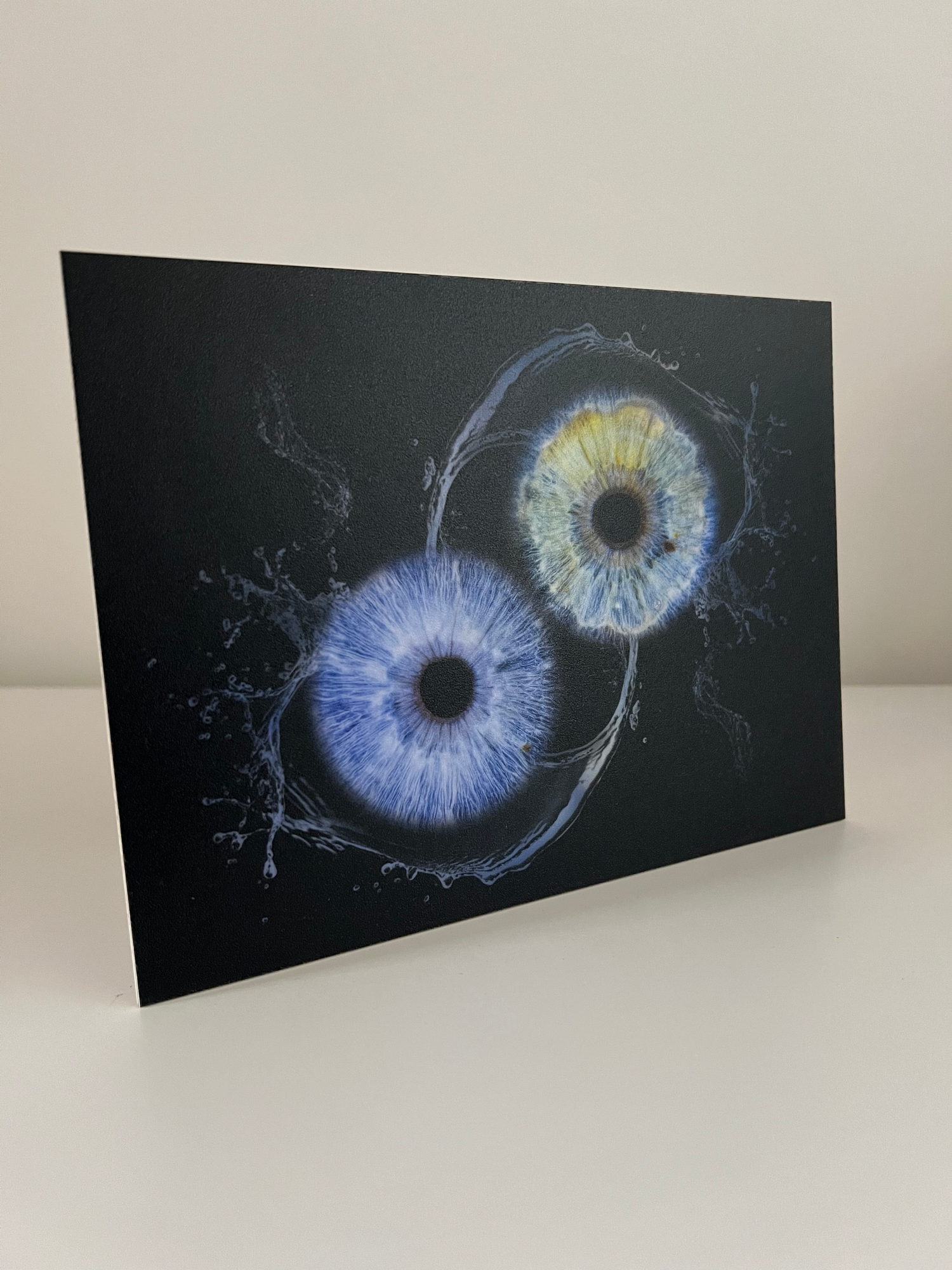 Artistic photograph of two detailed eye irises in blue and yellow with water splash effects on black background.