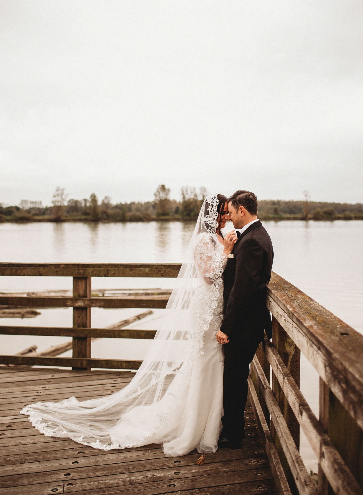 Wedding couple sharing an intimate moment on a waterfront dock, bride in a lace gown with a flowing cathedral veil.