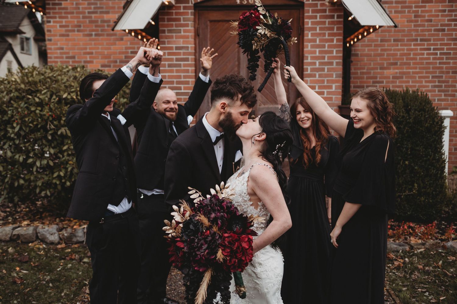 Bride and groom kiss surrounded by cheering wedding party outside a charming venue with string lights.