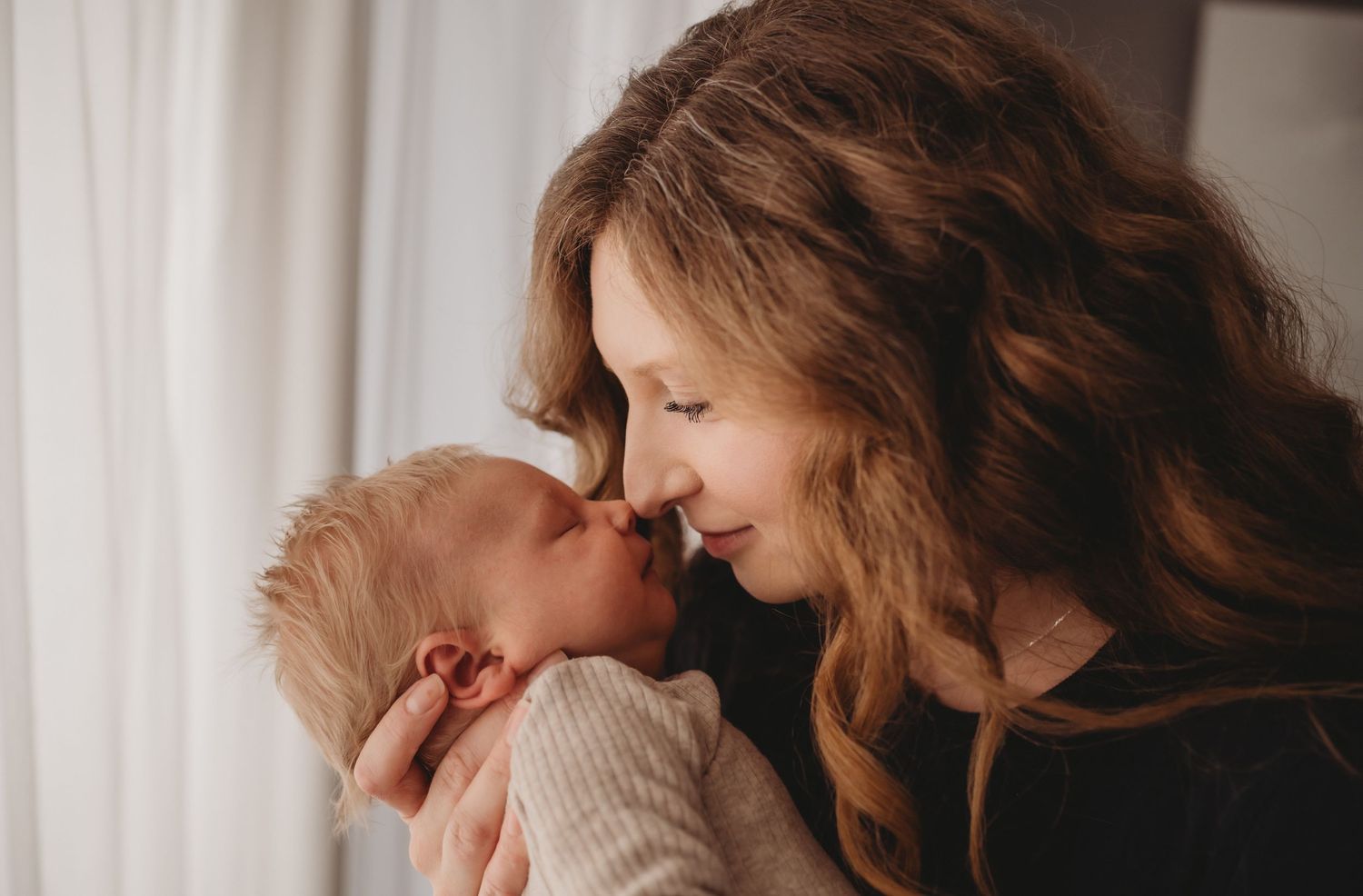 Tender moment of mother with long wavy hair smiling nose-to-nose with her newborn baby.