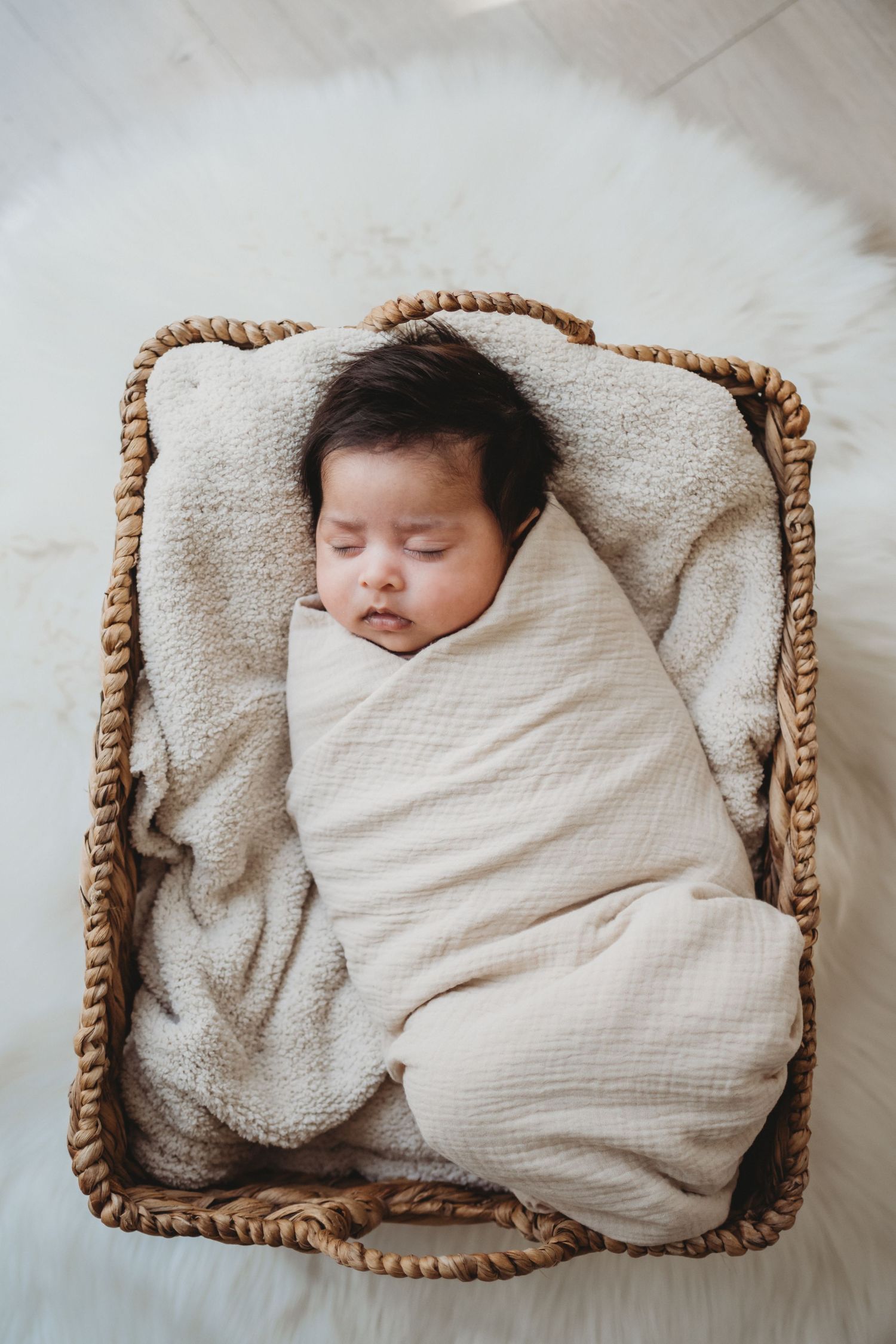 Newborn baby sleeping peacefully swaddled in a cream wrap inside a wicker basket on a white fur rug.