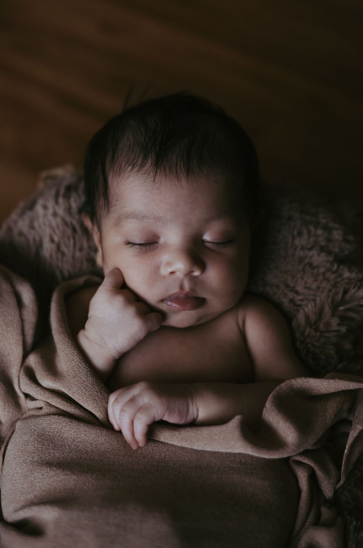 Sleeping newborn baby resting chin on hand wrapped in brown blanket in dark setting.