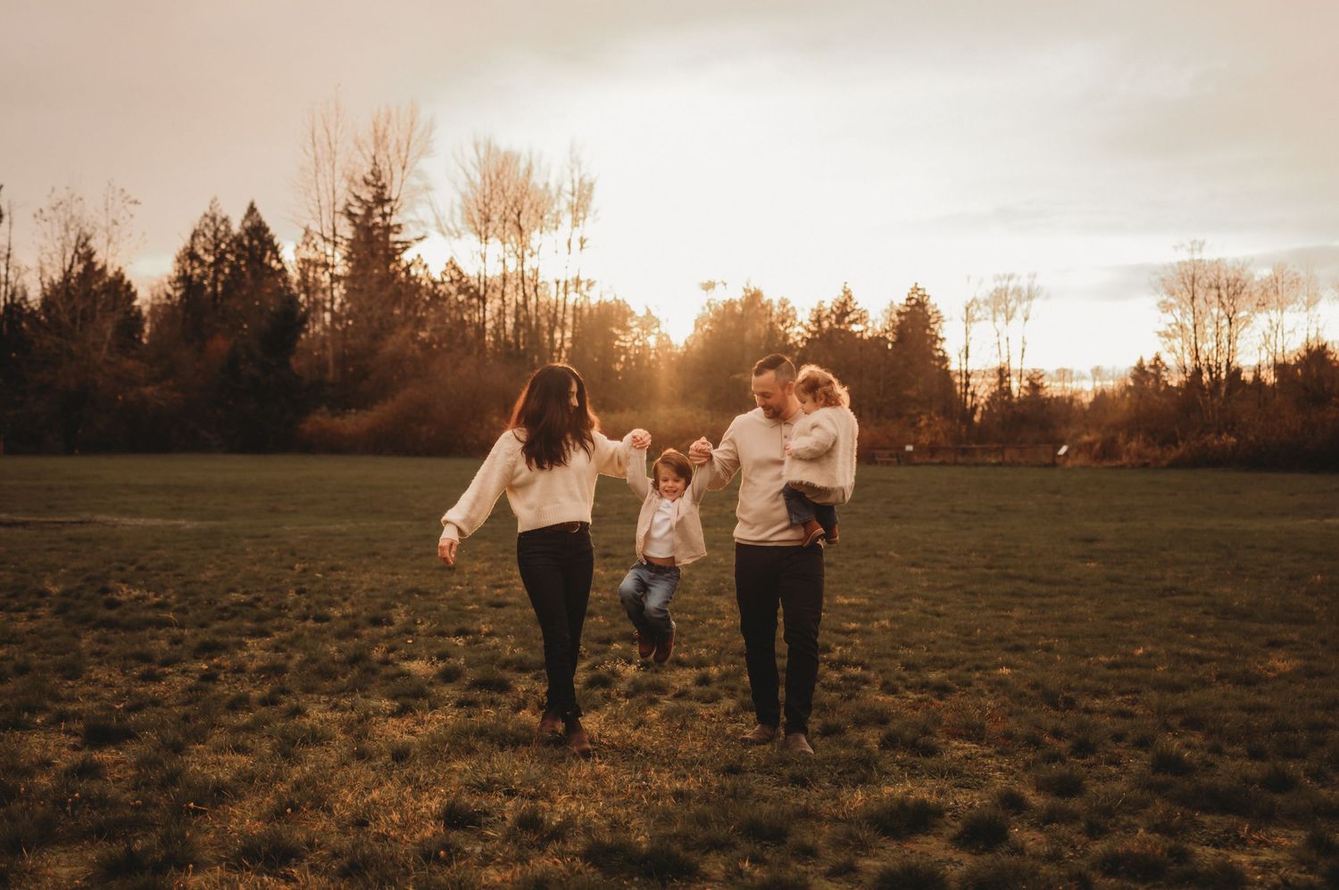 Family of four walking hand-in-hand across a sunlit green field at golden hour.
