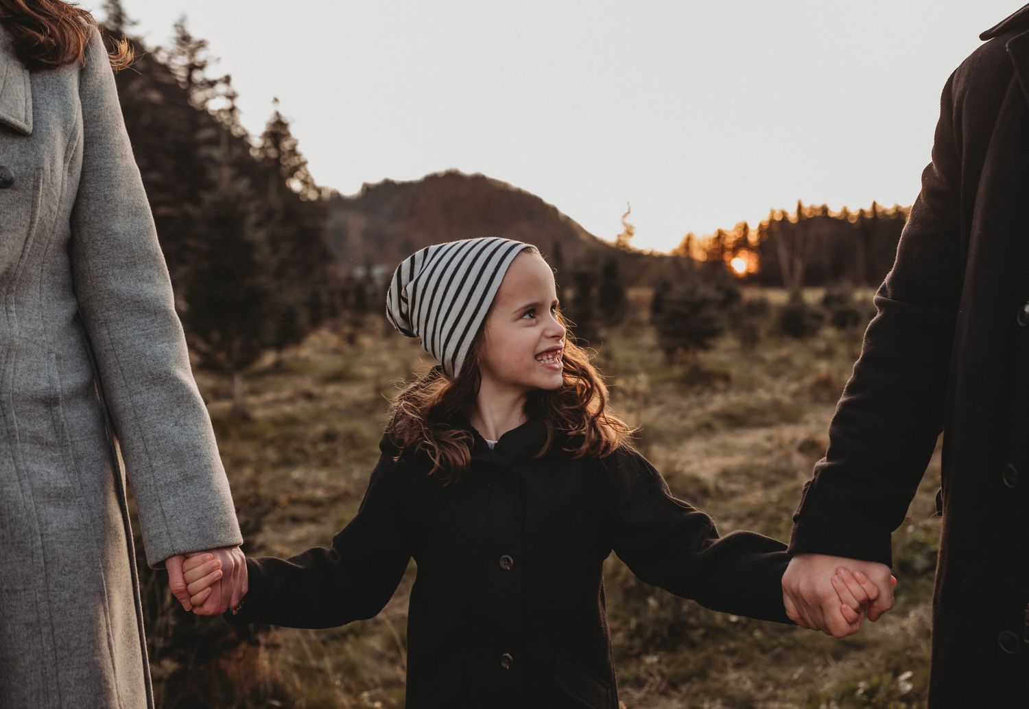 A happy child in a striped beanie holds parents hands walking through an autumn field at sunset.