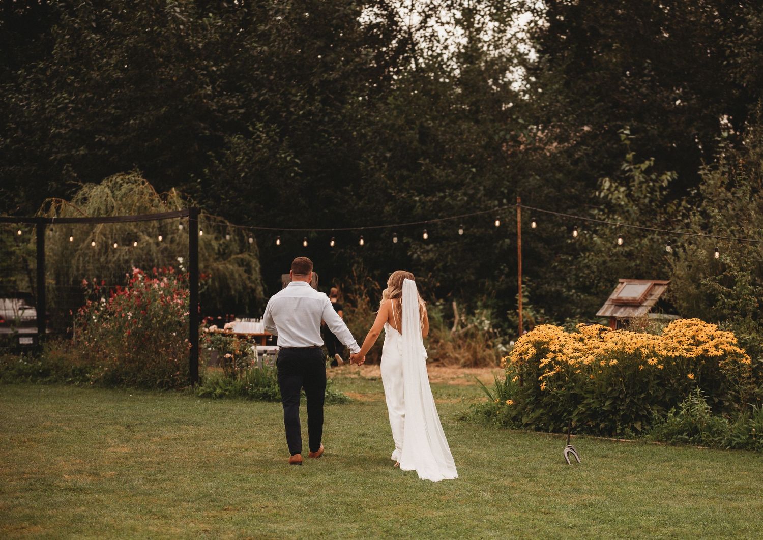 A bride and groom walk hand in hand through a lush garden venue decorated with string lights at dusk.