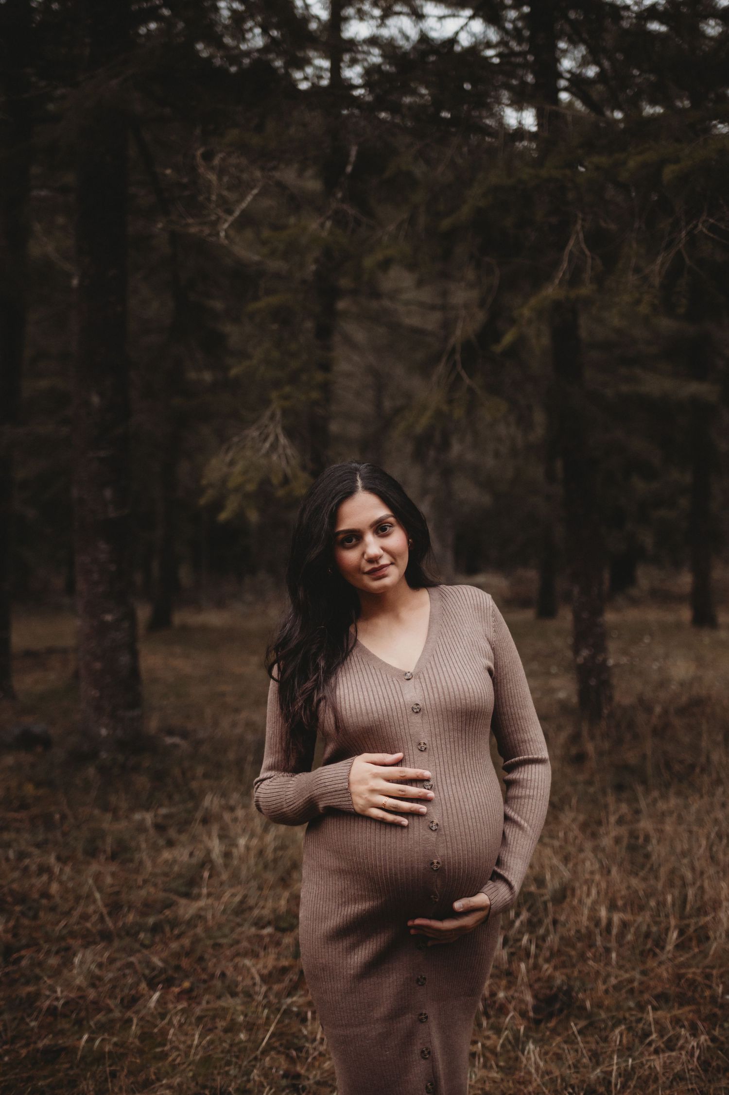 Smiling pregnant woman in brown ribbed dress holding baby bump in a dark forest.