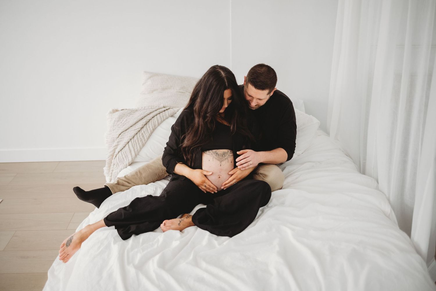 An expecting couple sits closely on a white bed, both lovingly touching the womans pregnant belly.