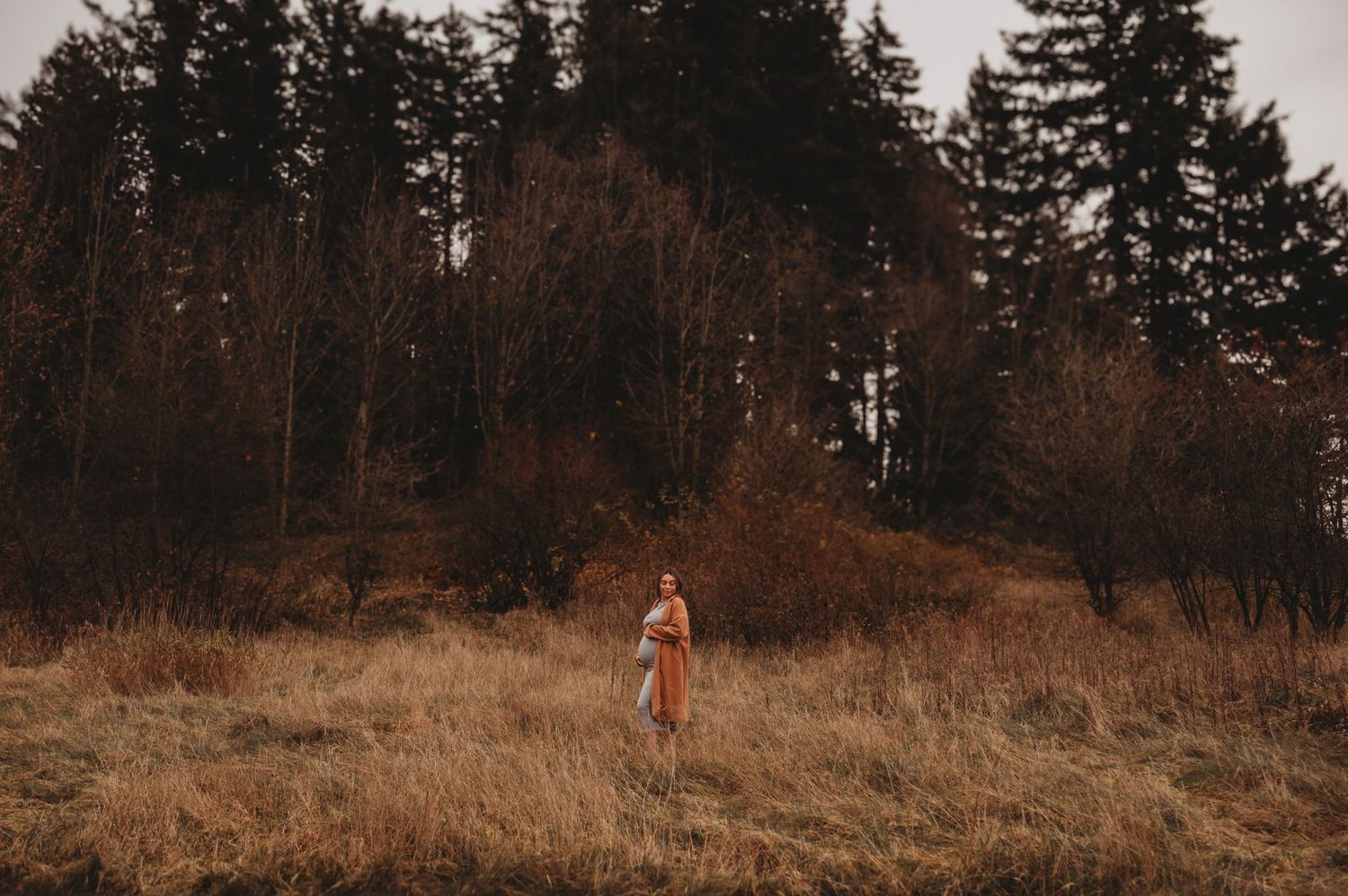 Pregnant woman in tan coat standing alone in autumn meadow.