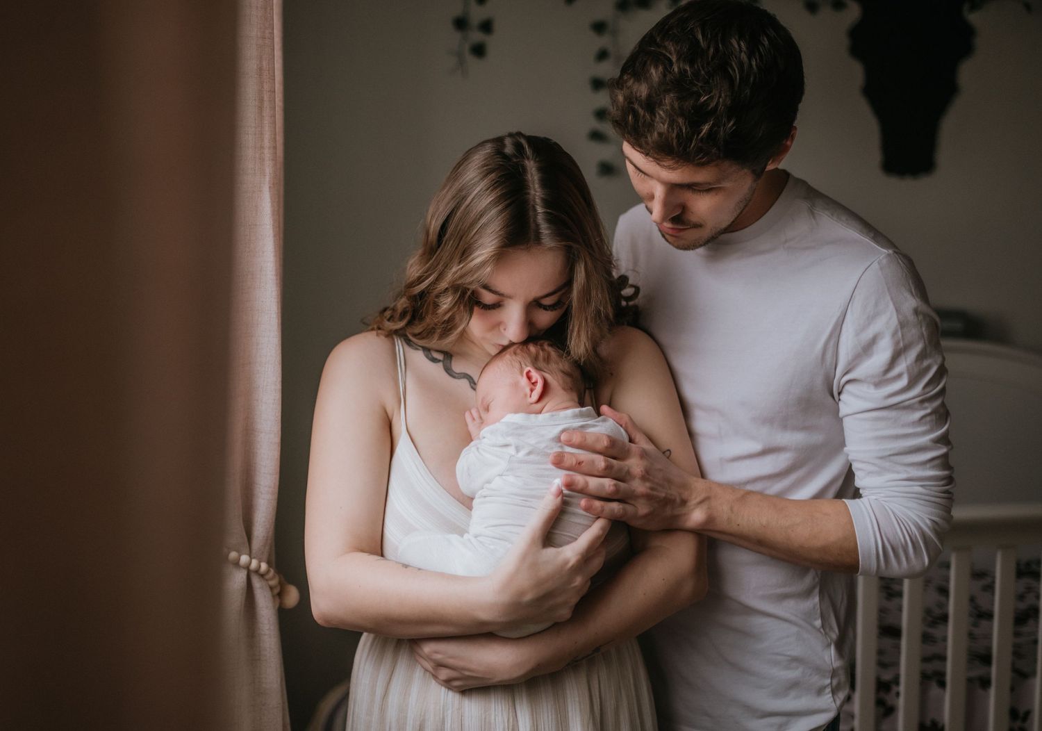 New parents tenderly cradling sleeping newborn baby wrapped in white swaddle cloth indoors.