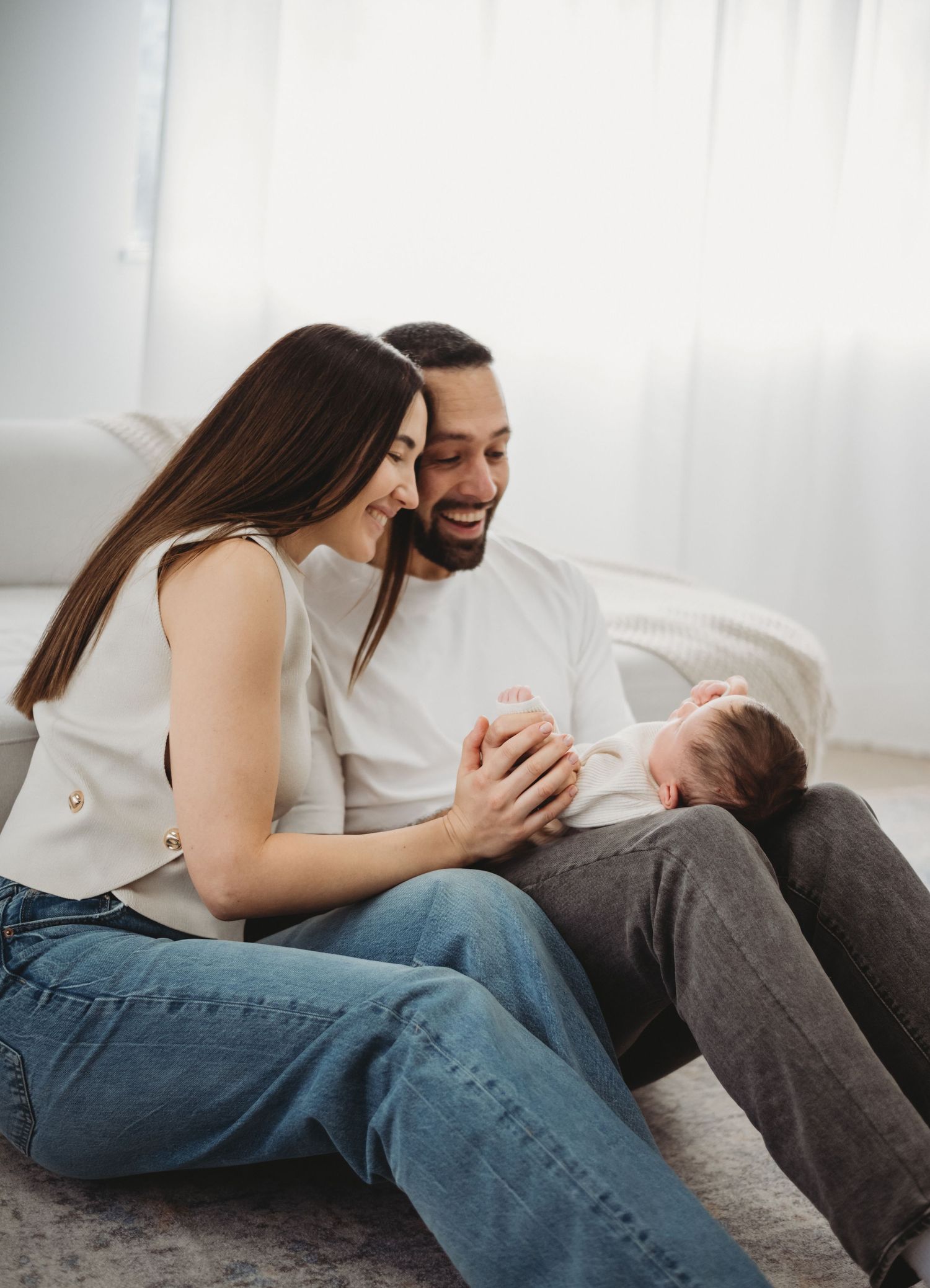 Happy young parents sitting on floor smiling at their newborn baby in a bright white room.