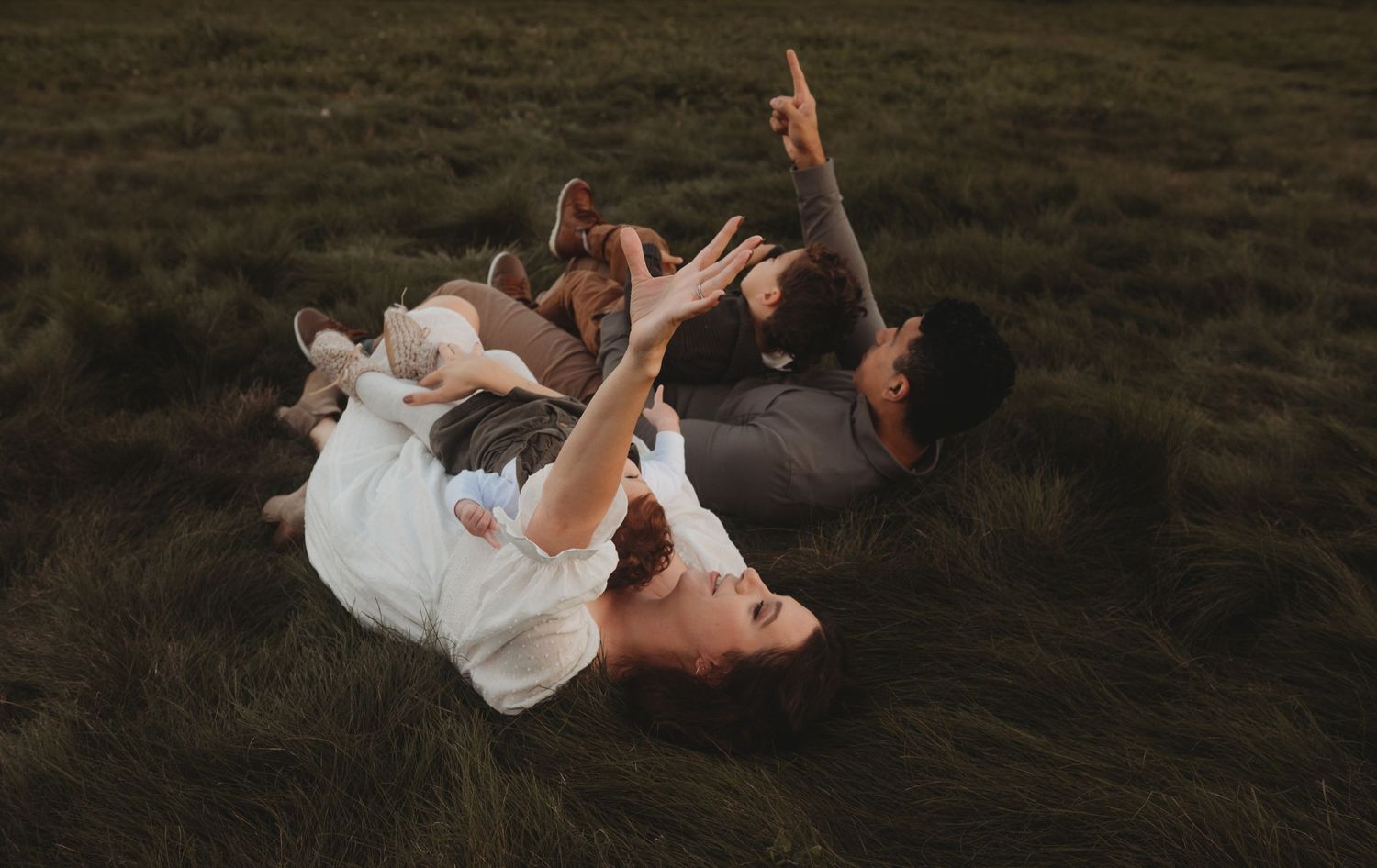 A candid family photo of parents and two children lying on green grass, arms raised in playful joy.