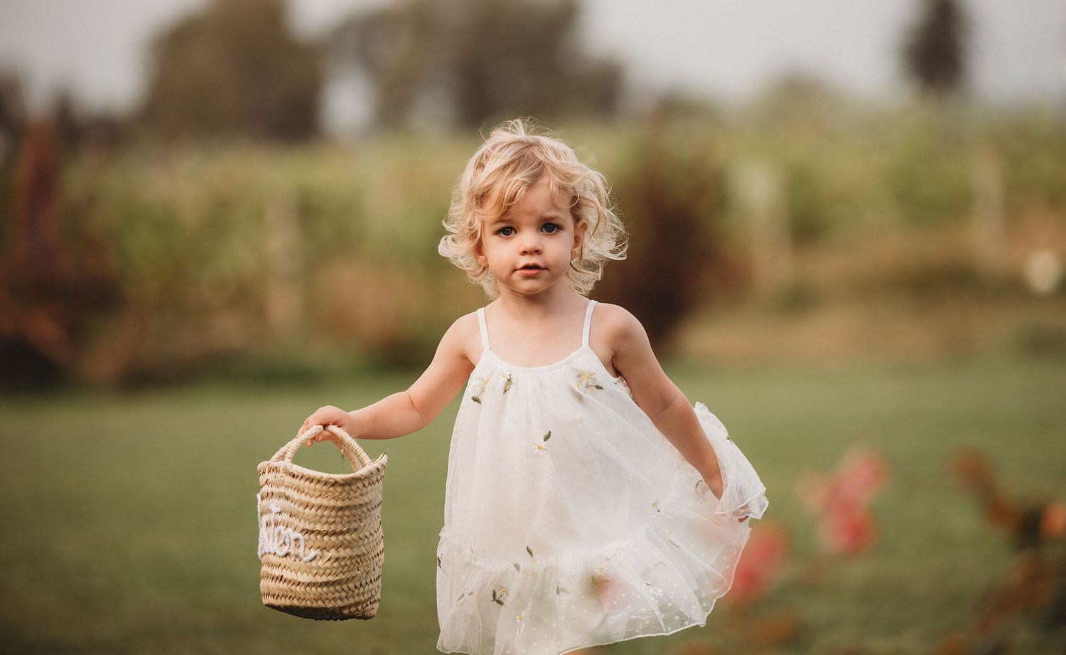 Adorable toddler girl in white dress carries a wicker basket outdoors.