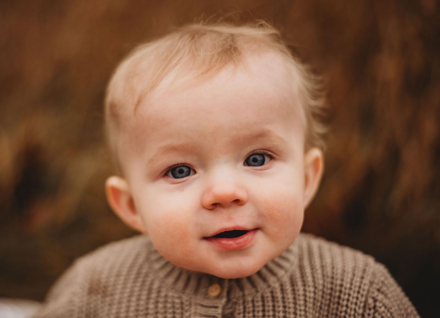 An adorable blue-eyed baby in a brown knit sweater smiles at the camera with a warm bokeh background.