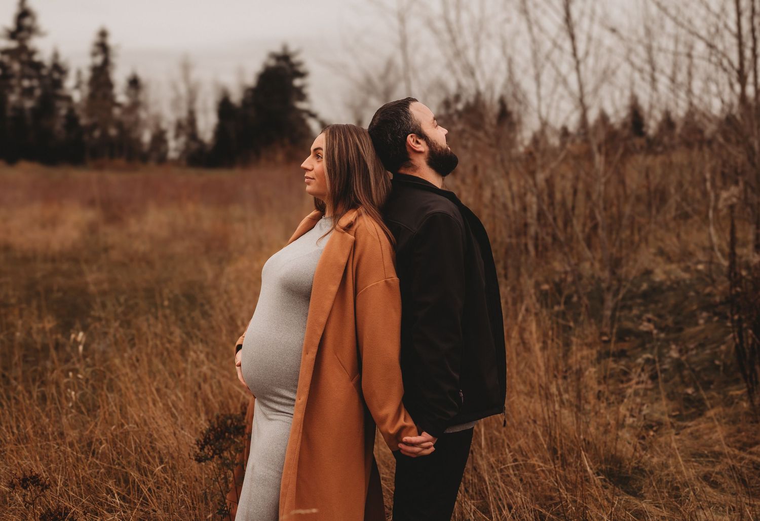 Pregnant couple standing back-to-back holding hands in an autumn field at dusk.