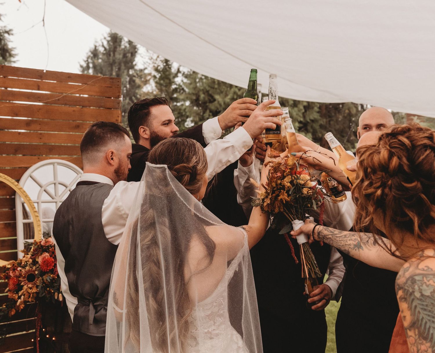 Wedding party raises beer bottles in a celebratory toast under a white tent outdoors.