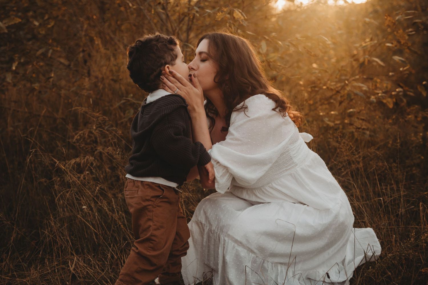Mother and young son sharing a sweet kiss outdoors in warm golden sunset light among tall grasses.
