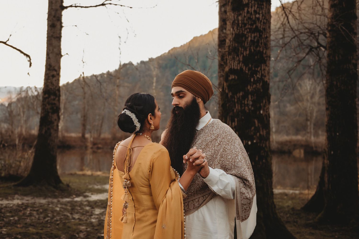 Sikh groom with long beard holds bride's hands tenderly in a forest with mountain backdrop.