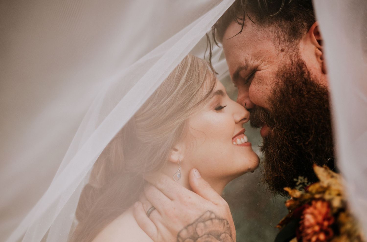 Romantic bride and bearded groom share a tender moment beneath flowing white wedding veil.