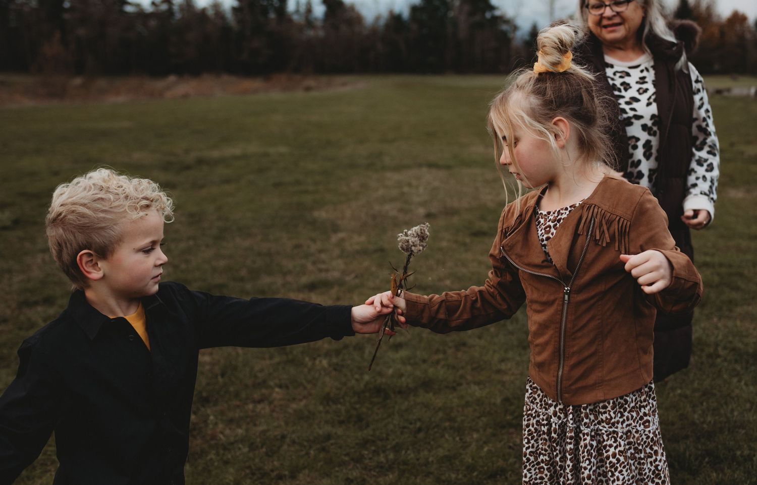 A young boy and girl share a wildflower in an open green field while another child watches nearby.
