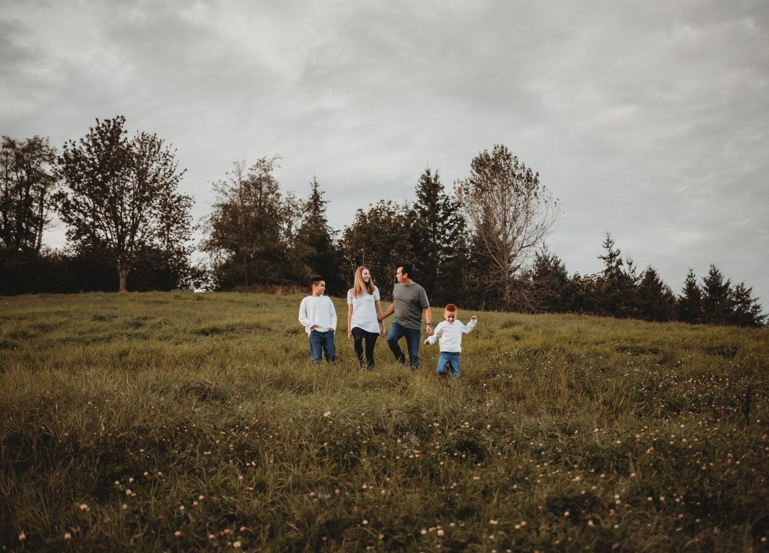 A family of four walks hand in hand through a wide open grassy field under cloudy skies.