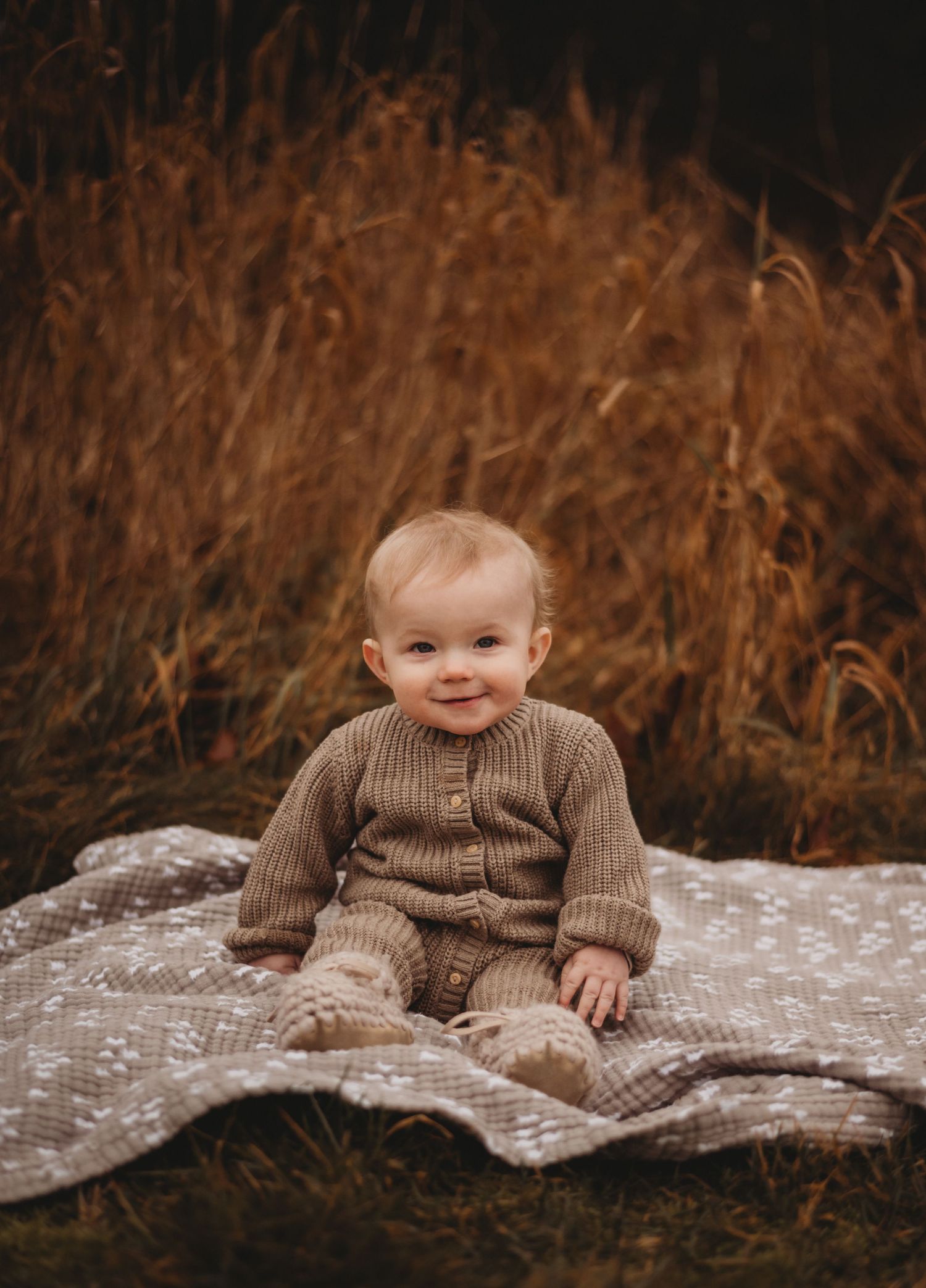 Adorable baby in brown knit outfit sitting on blanket in golden autumn grass field.