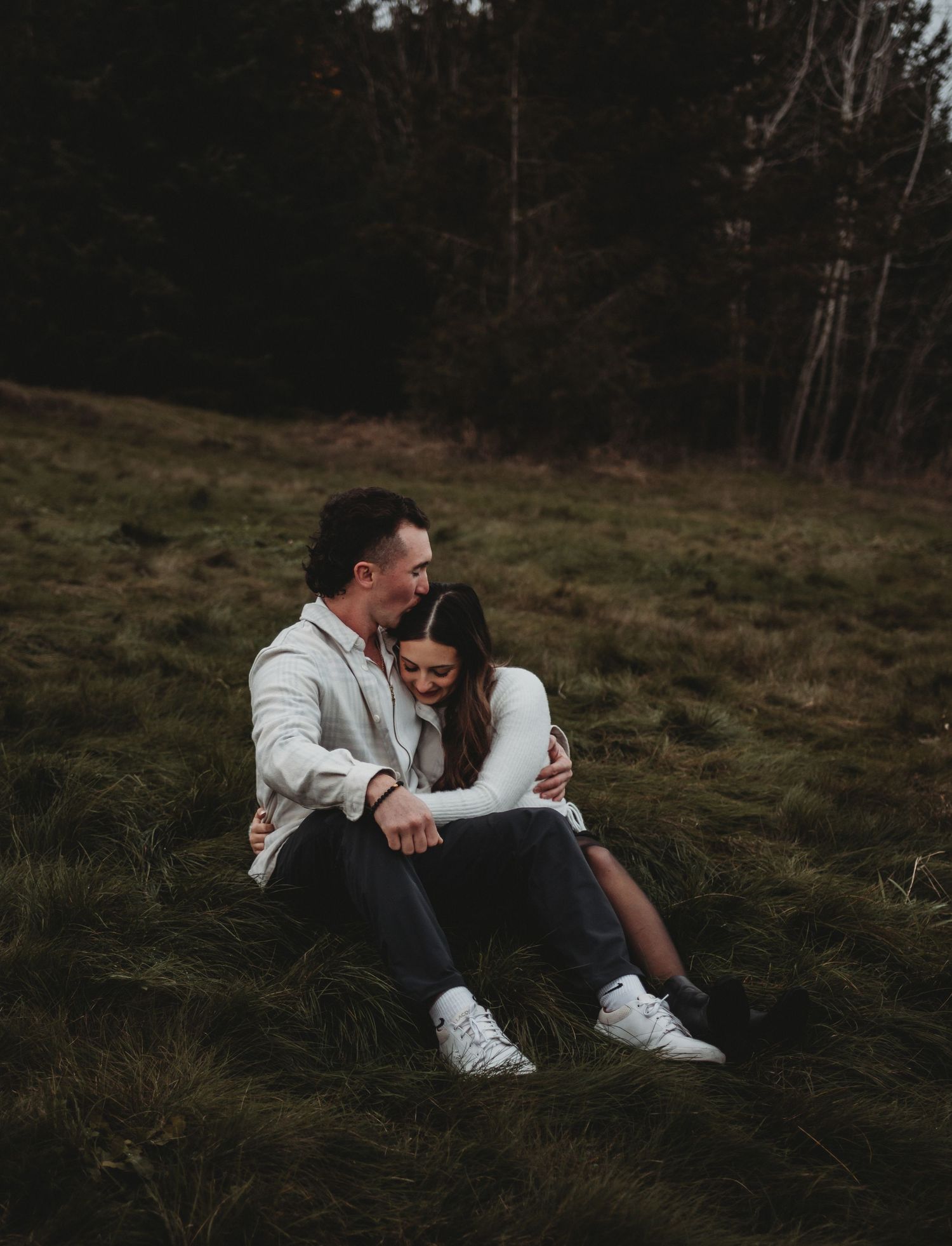 Couple sitting together on a grassy field, sharing a tender embrace in soft moody light.