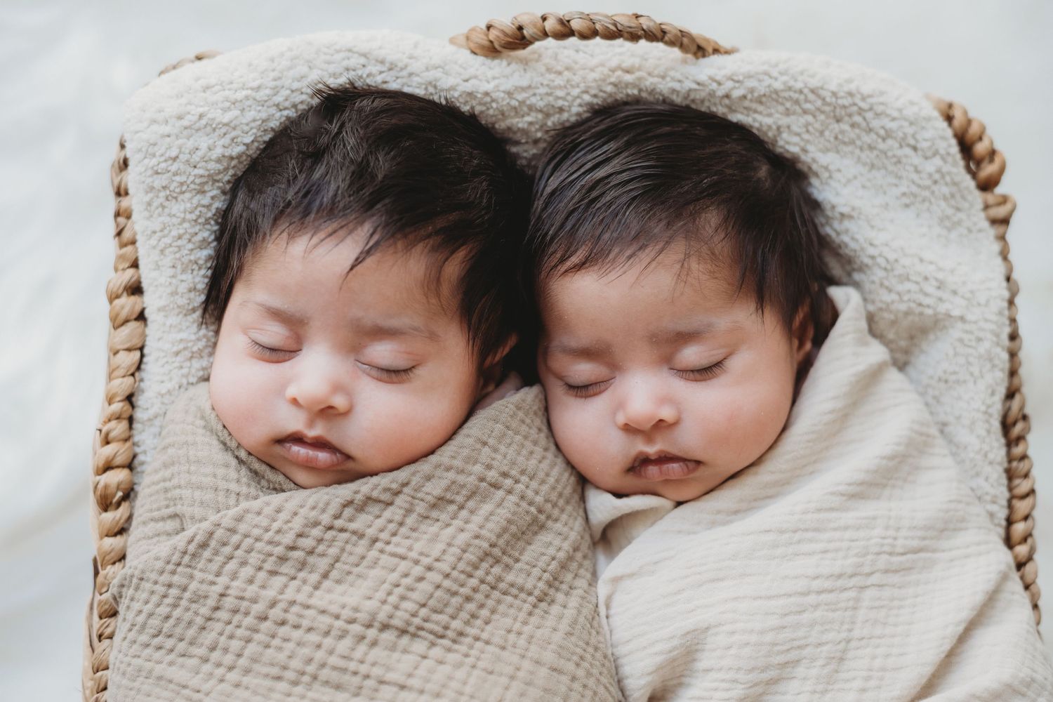 Twin newborn babies sleeping together swaddled in cream muslin wraps inside a wicker basket.