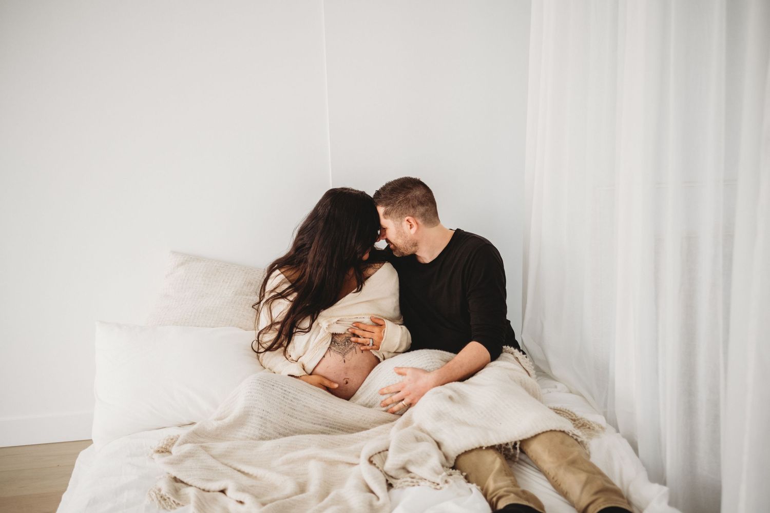 Couple sharing intimate moment on white bed during maternity photoshoot.