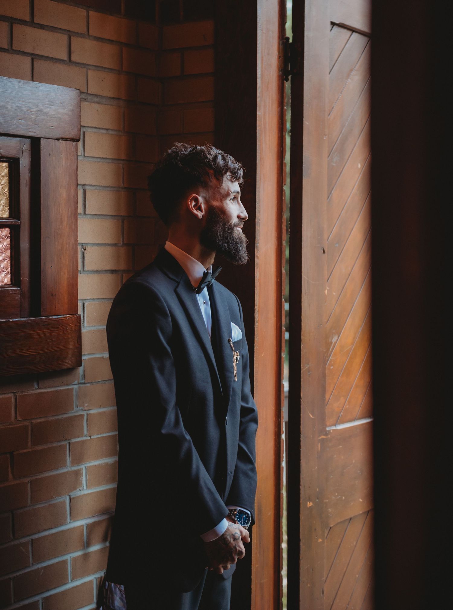 Bearded groom in navy suit gazing thoughtfully through wooden church doors on wedding day.
