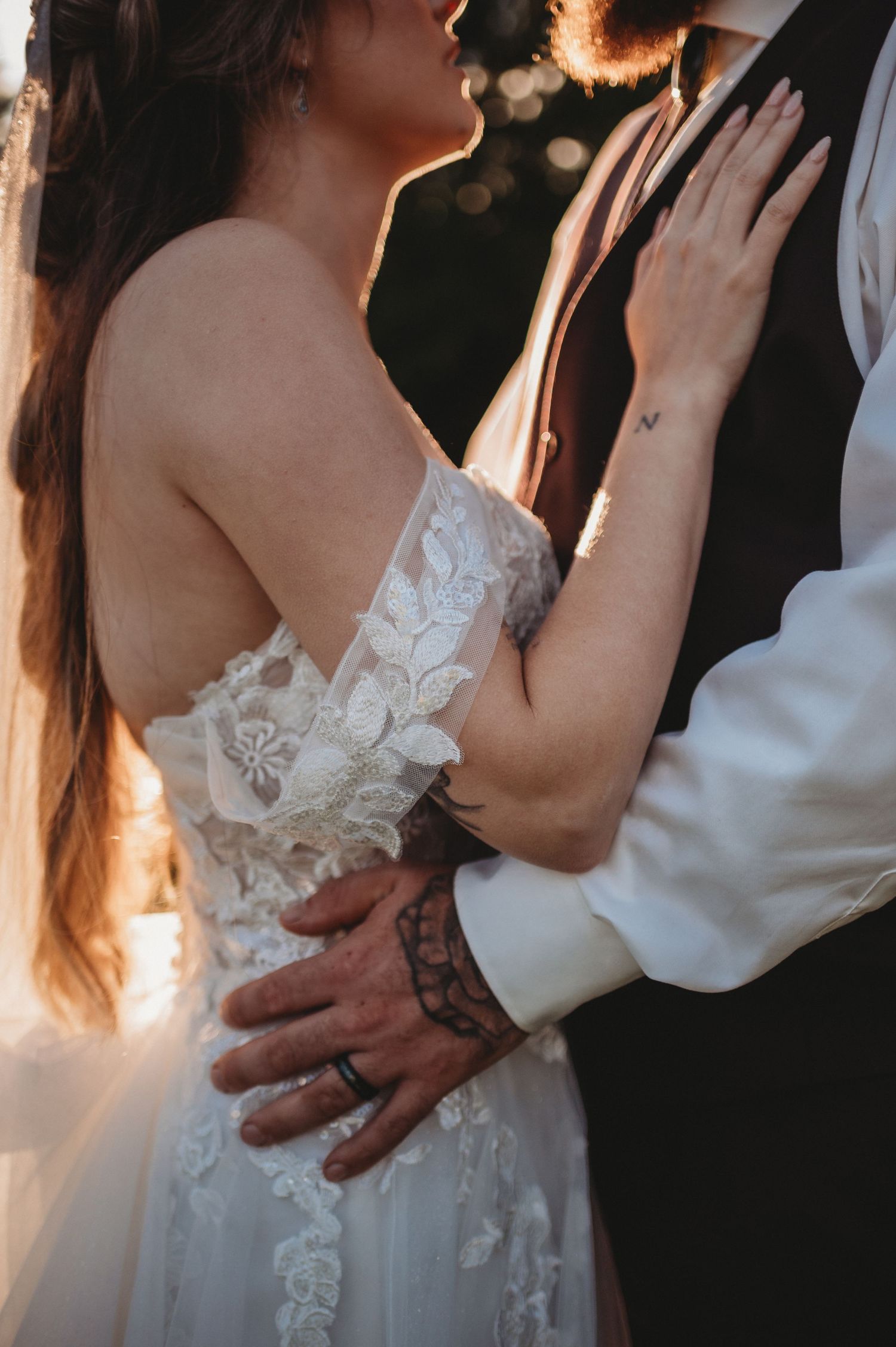 Close-up of bride and groom embracing, showcasing intricate lace wedding gown back and groom's tattoo sleeve.