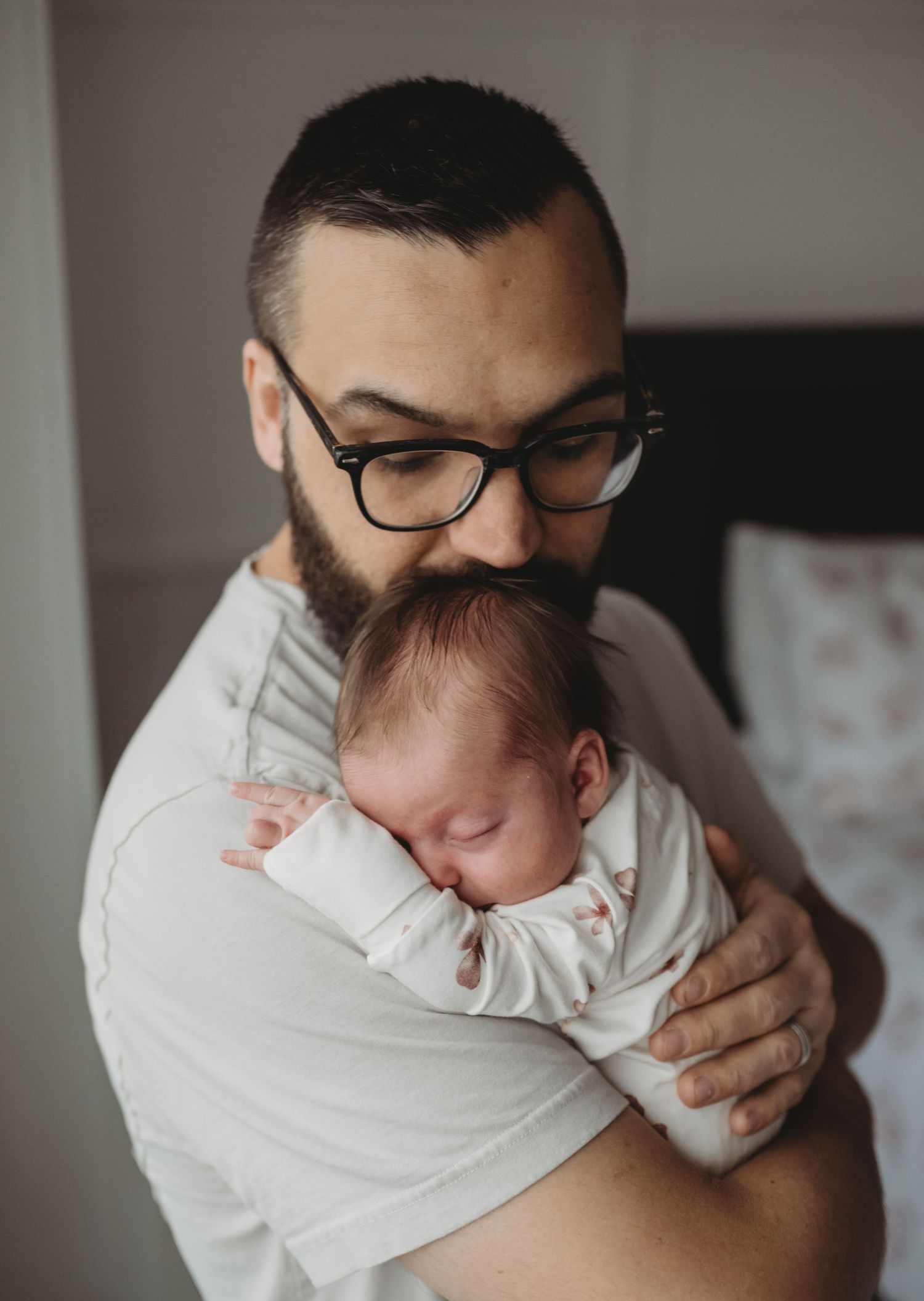 A bearded father with glasses lovingly cradles his sleeping newborn against his shoulder.