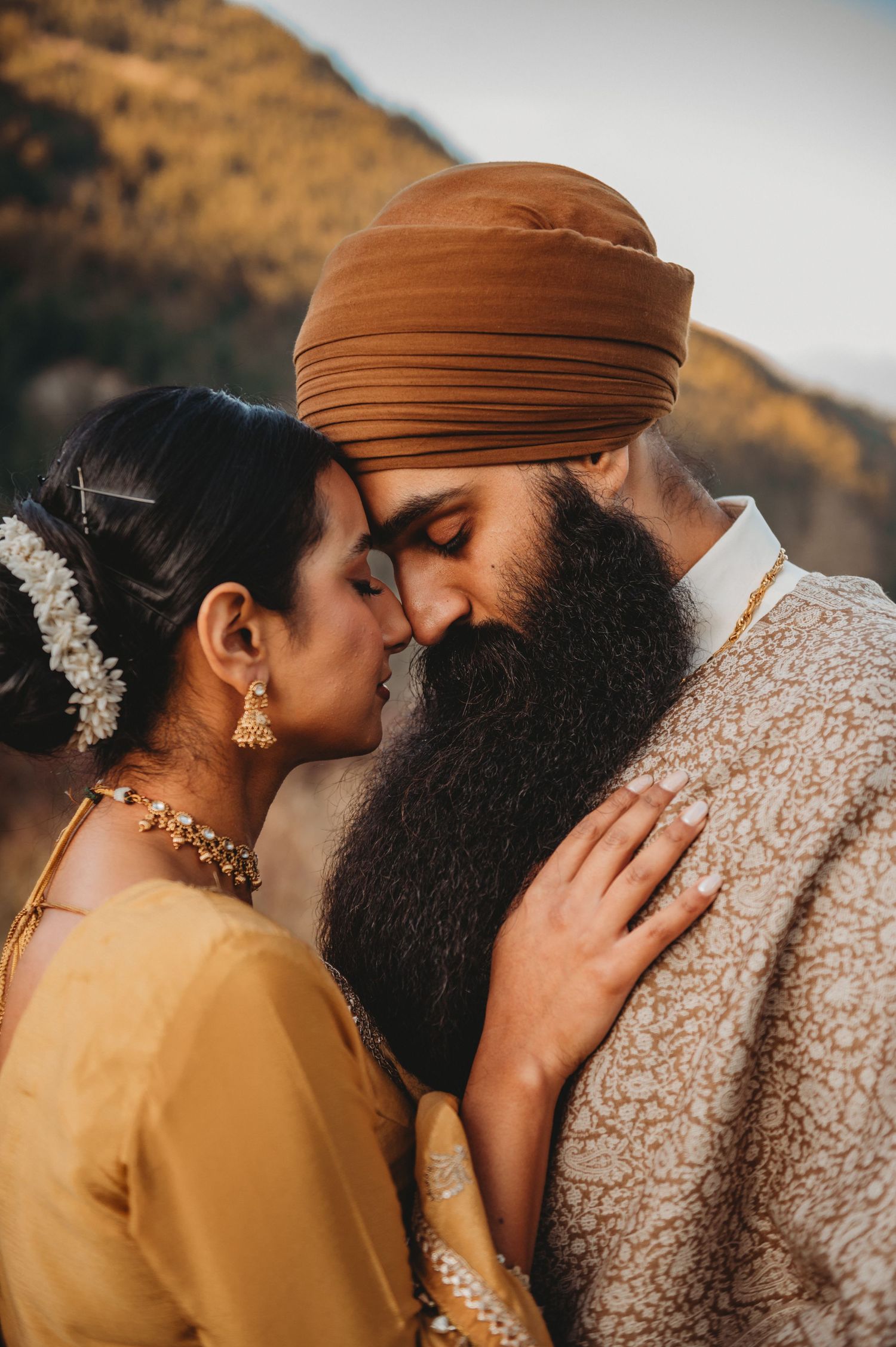 Sikh couple in traditional attire sharing an intimate moment outdoors, bride in gold jewelry and groom in brown turban.