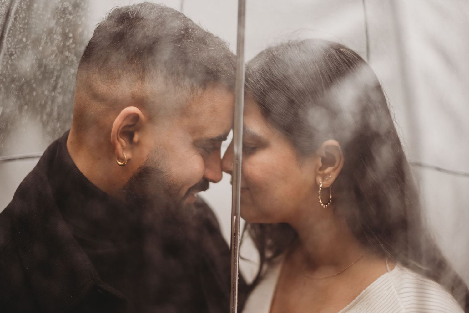 Couple gazing at each other closely through a rain-covered transparent umbrella.