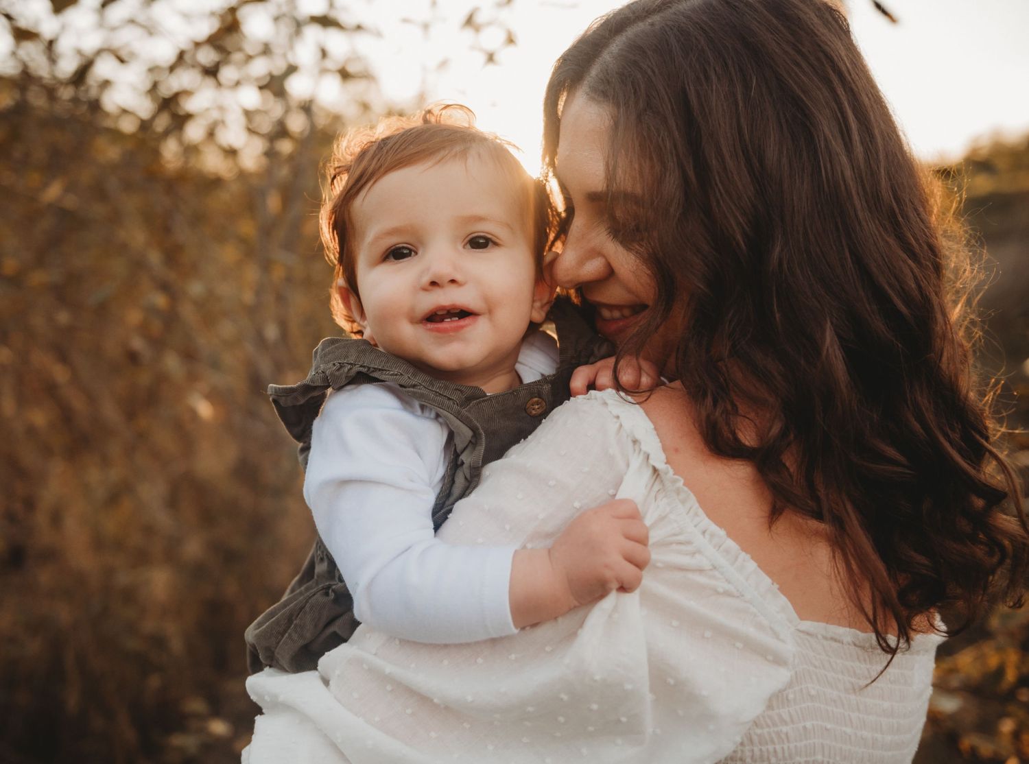 Smiling baby held by mother in warm fall sunlight.