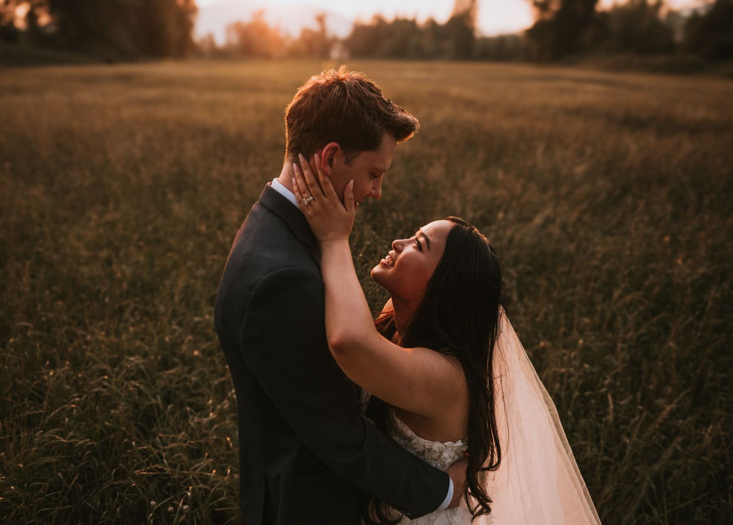 Bride tenderly holding groom's face in sunlit field during romantic outdoor wedding portrait.