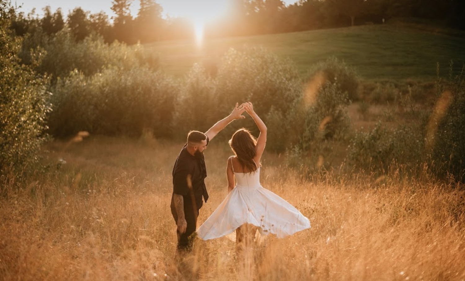 A bride in a white dress twirls joyfully with her groom in a sunlit golden field at sunset.