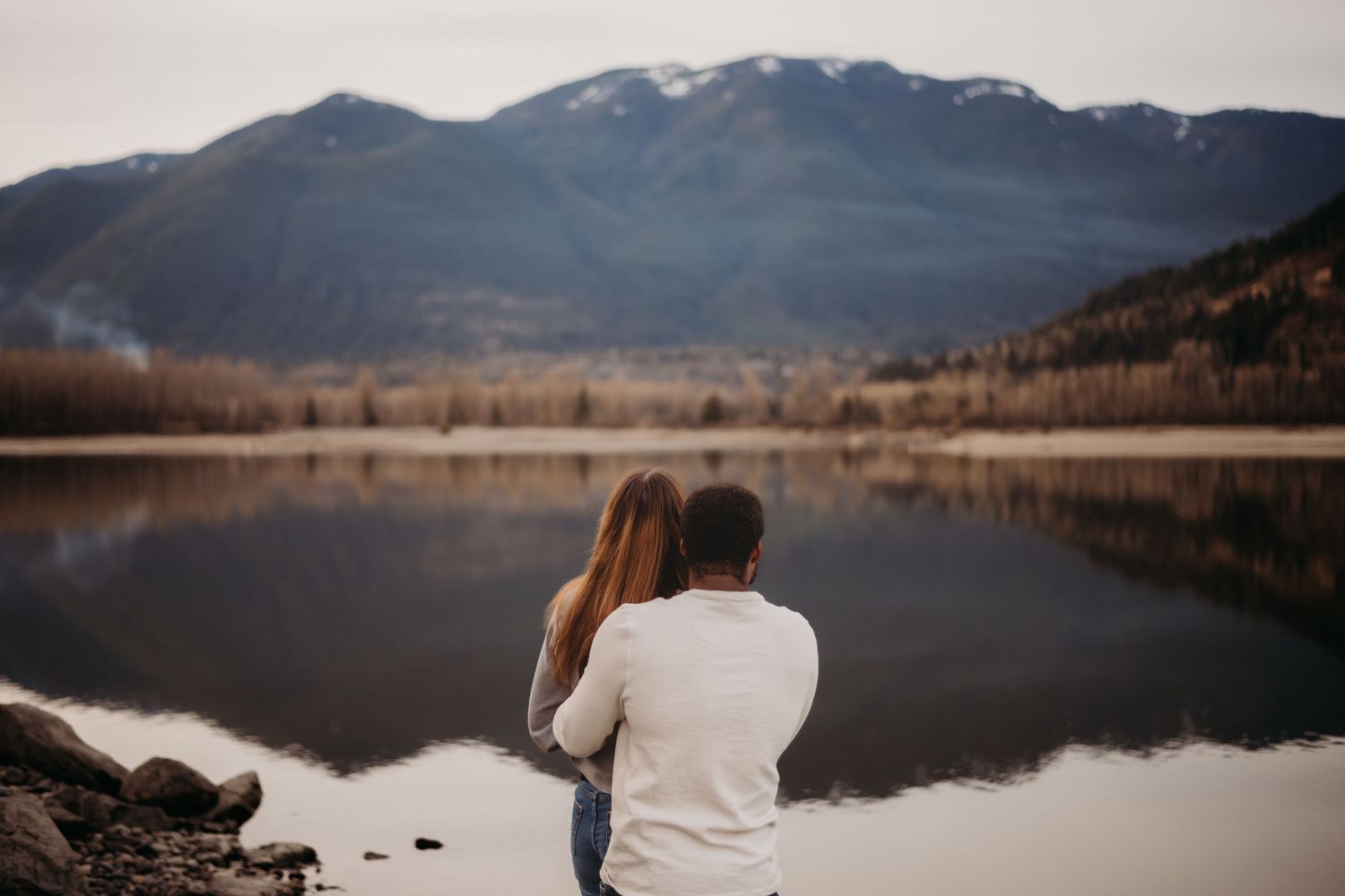 A couple sits together gazing at a stunning mountain lake reflection on a calm overcast autumn day.