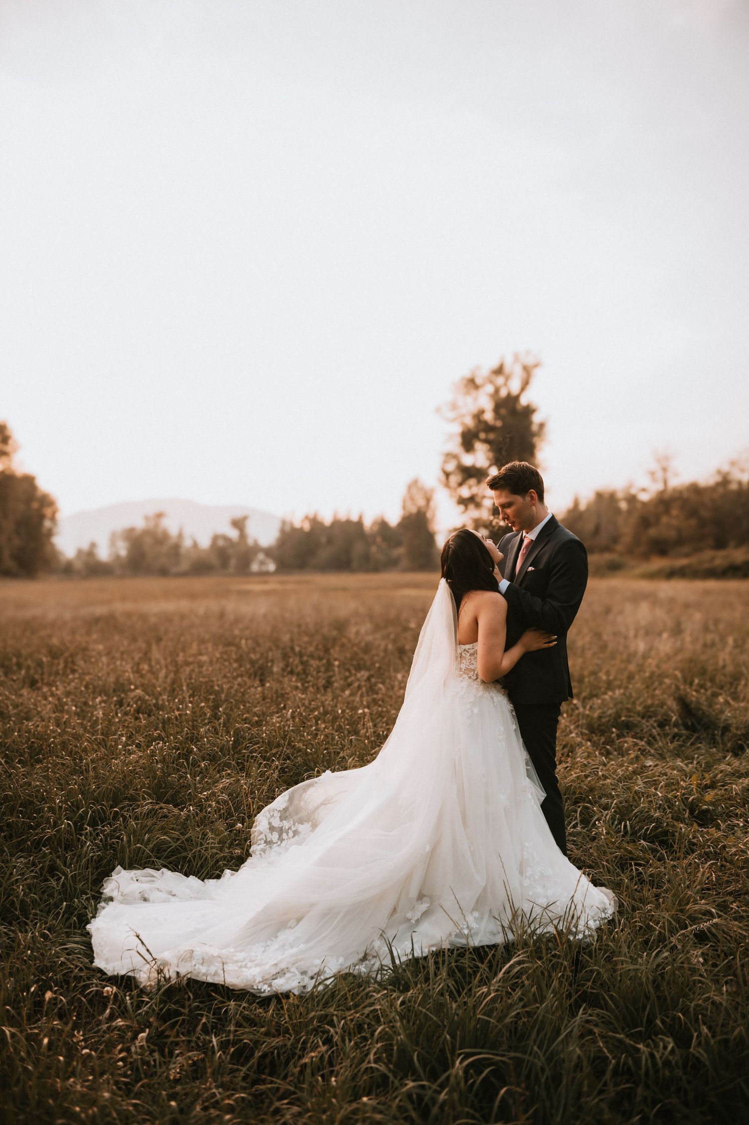 A bride and groom embrace in a golden field at sunset wearing elegant wedding attire.