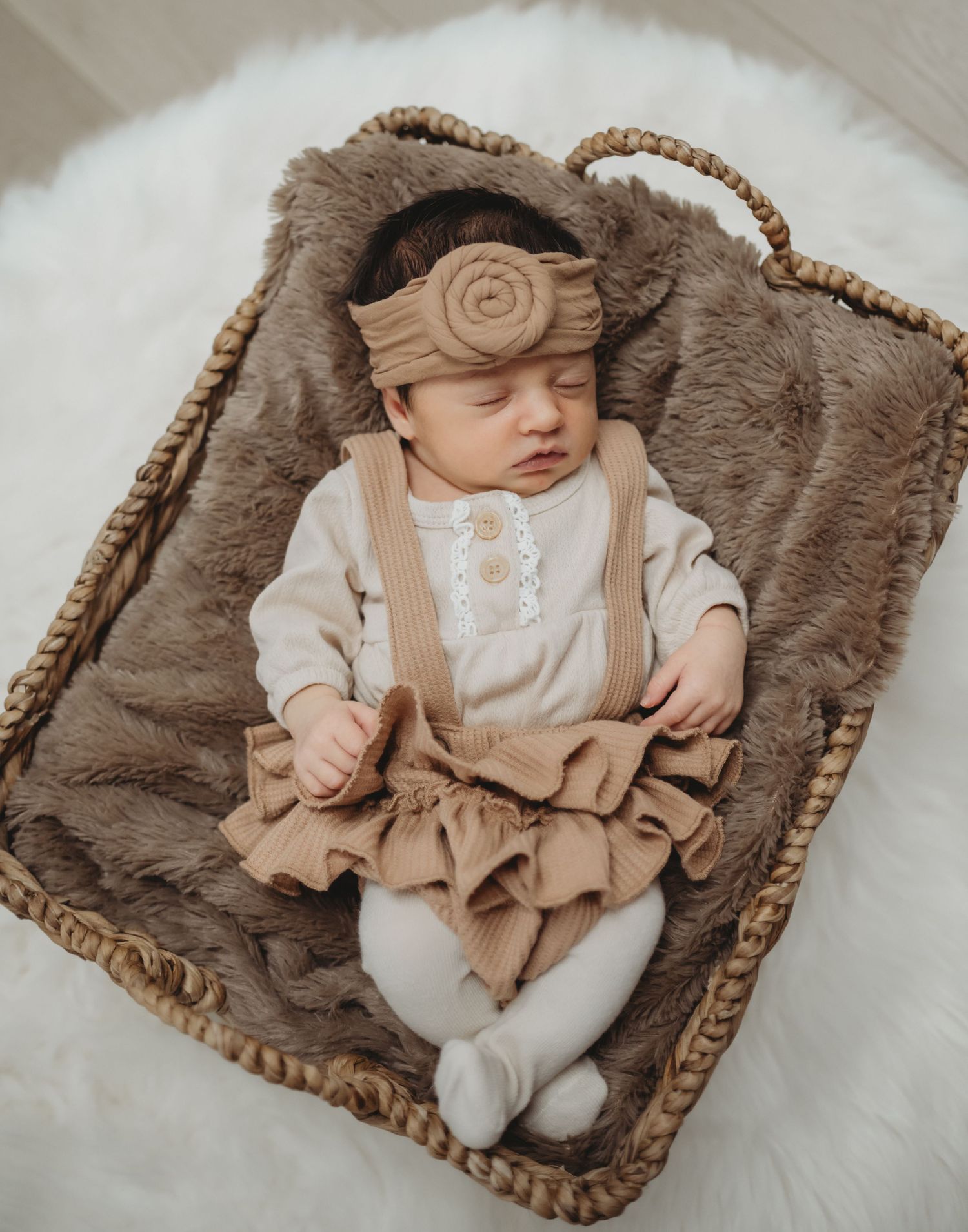 Sleeping newborn baby girl in ruffled tan dress and headband nestled in wicker basket with fur blanket.