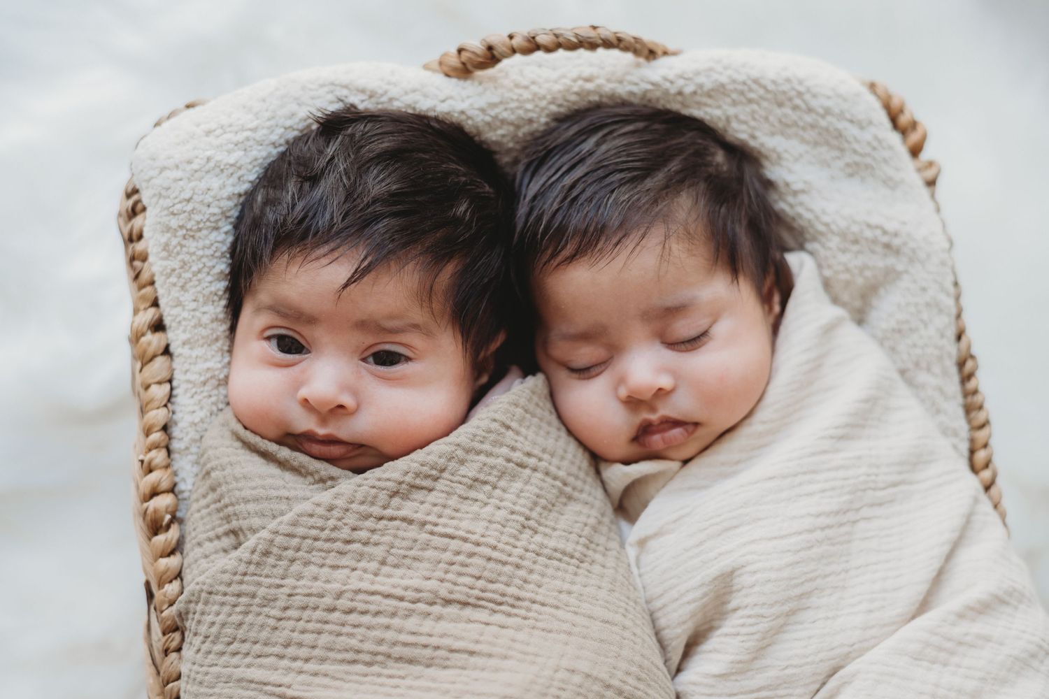 Twin newborns swaddled in cream wraps in a wicker basket, one awake and one sleeping peacefully.