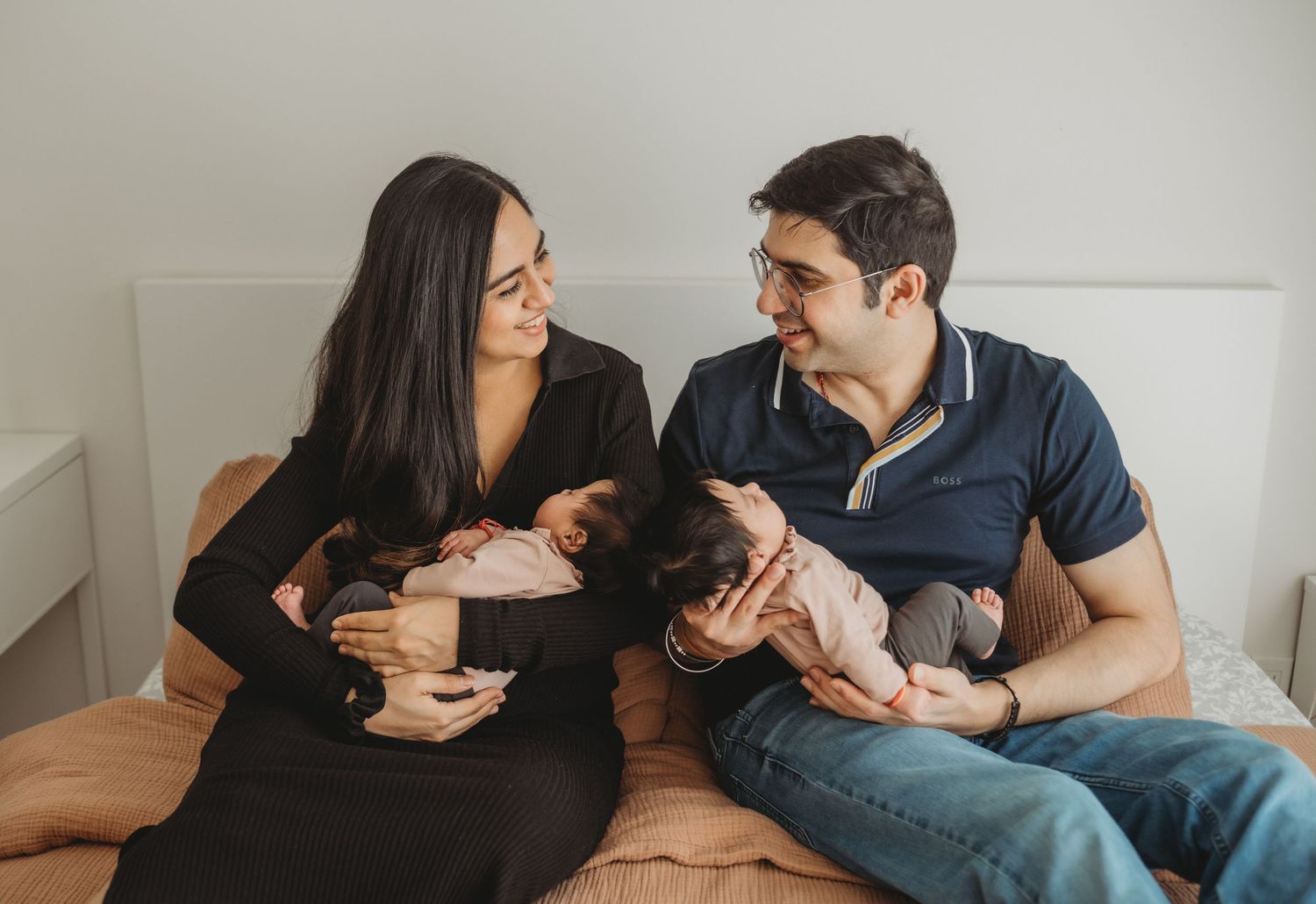 Happy parents sitting on bed holding newborn twins, smiling at each other in bright bedroom.